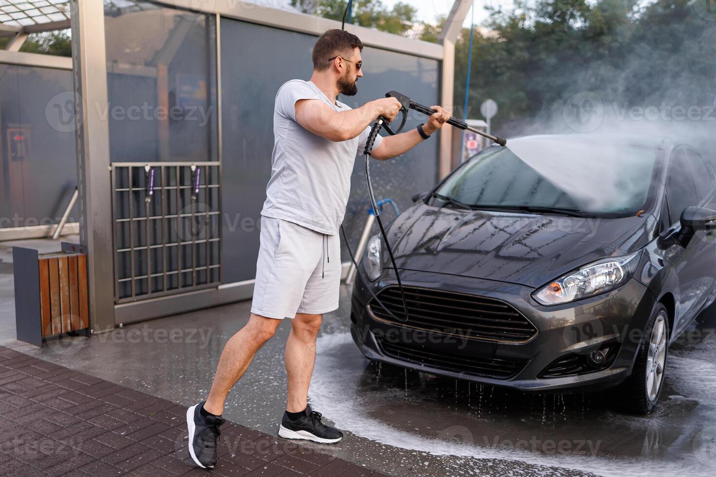 A man stands in front of the car and points a water cannon at it, washing the water off of it. A car at a self service car wash. photo