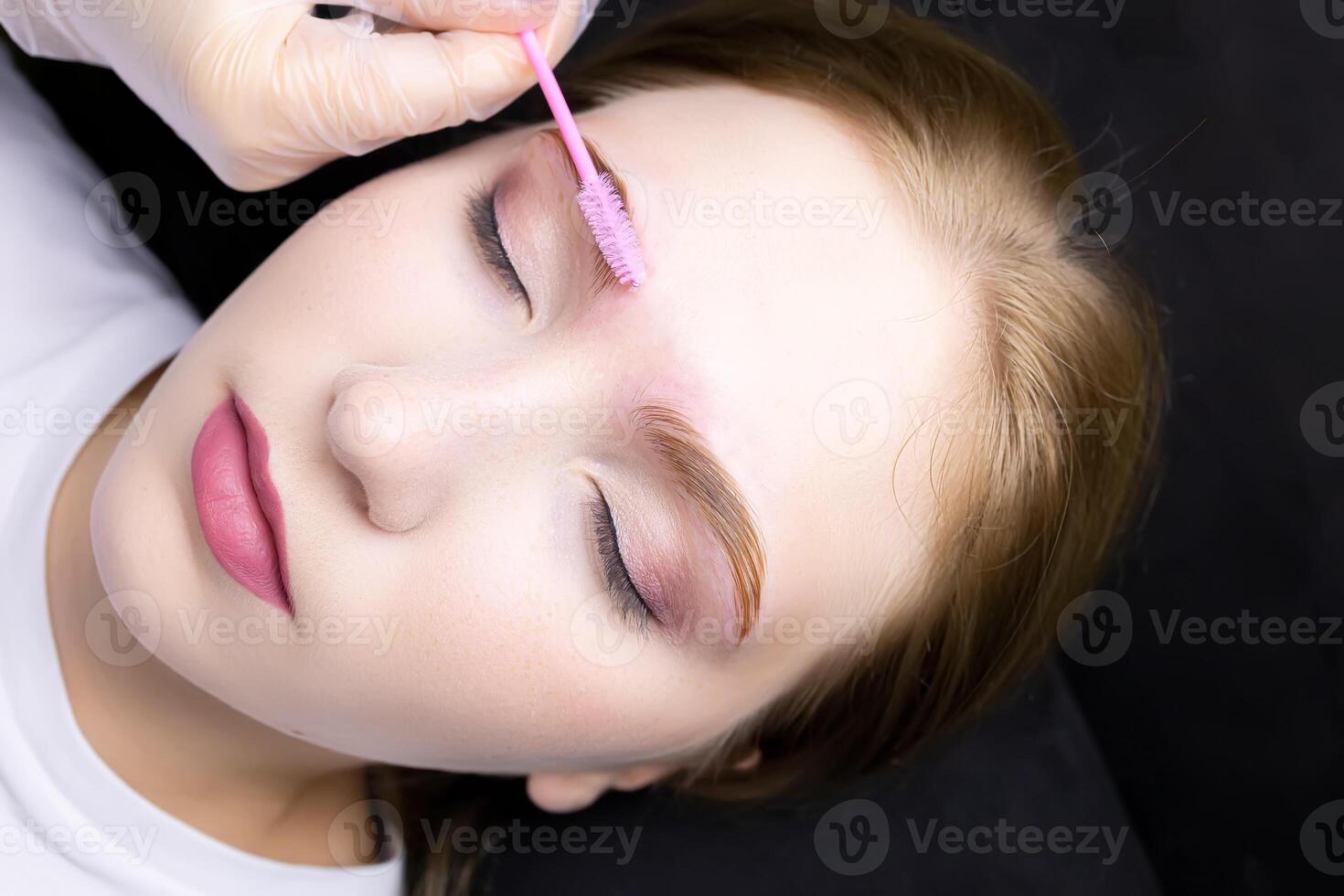 a close-up of the face of a blonde model who lies on the eyebrow lamination procedure, the master combs the eyebrows with a brush photo