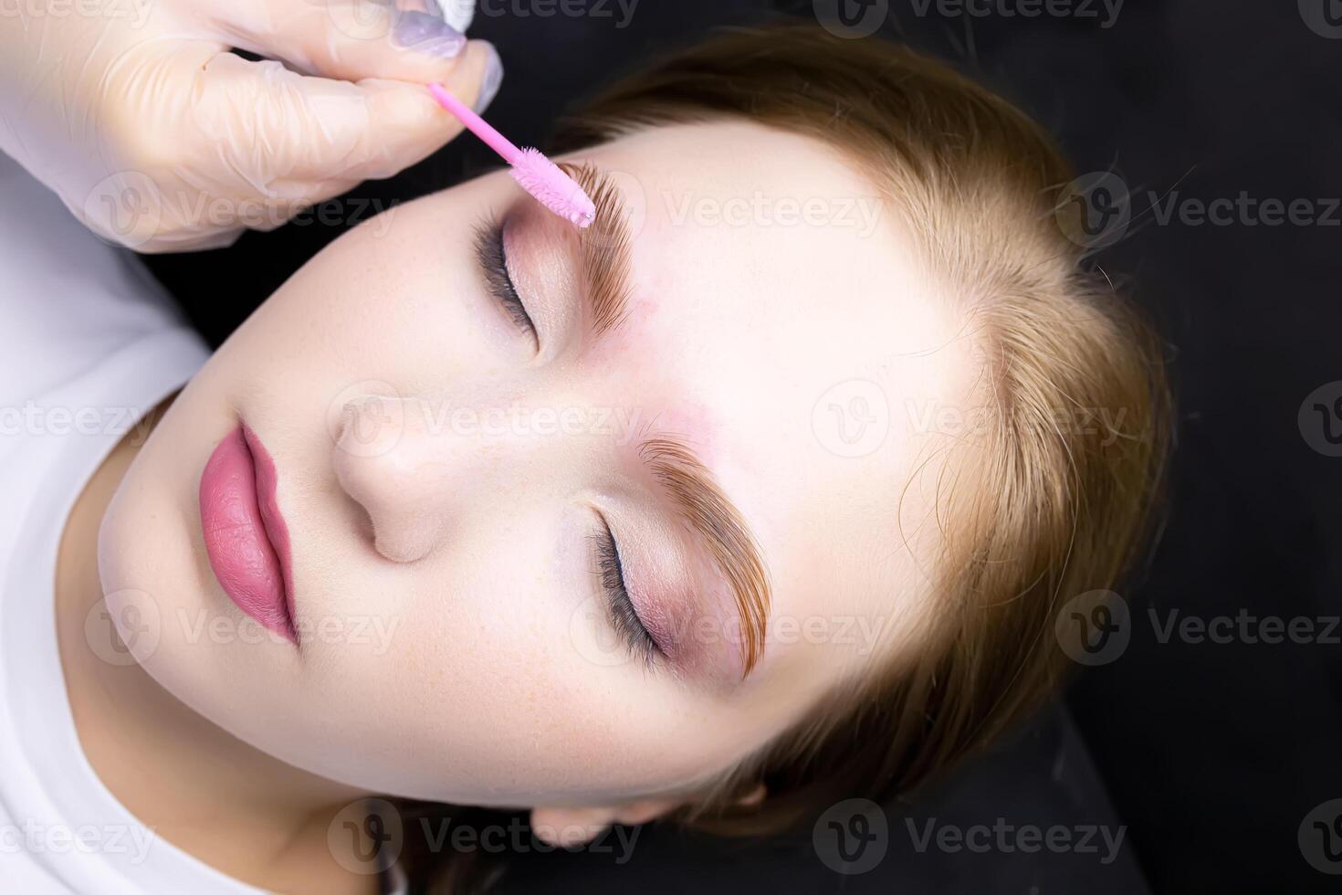 a close-up of the face of a blonde model who lies on the eyebrow lamination procedure, the master combs the eyebrows with a brush photo