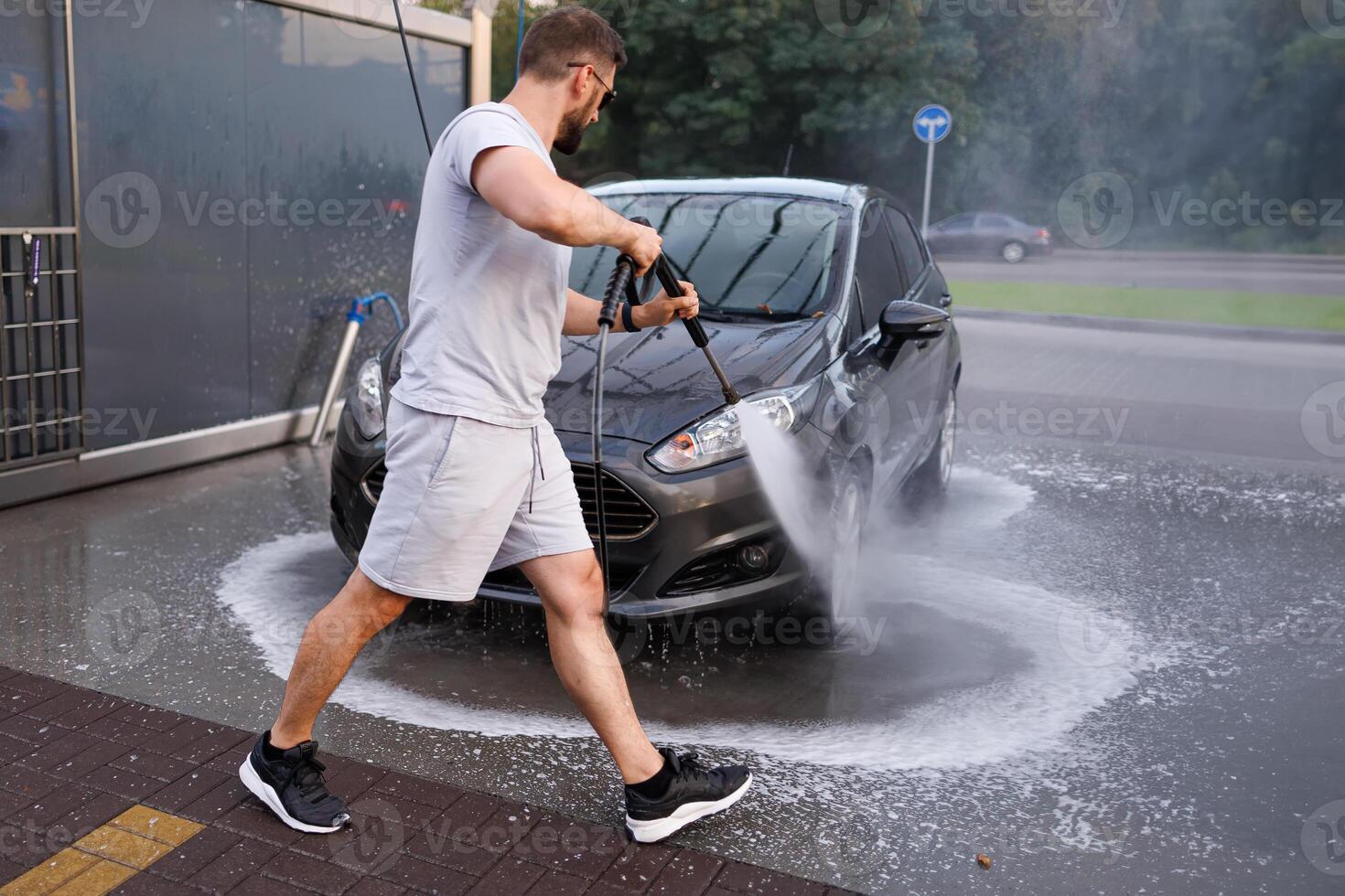 A man stands in front of the car and points a water cannon at it, washing the water off of it. A car at a self service car wash. photo