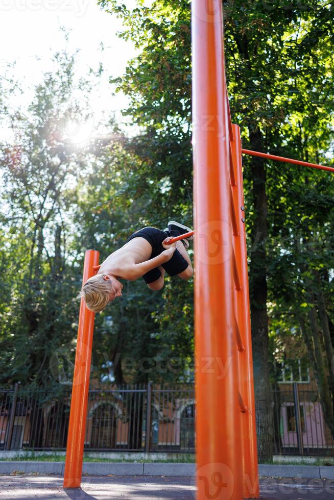 A teenager in a park playing sports hangs upside down on a crossbar. Street workout on a horizontal bar in the school park. photo