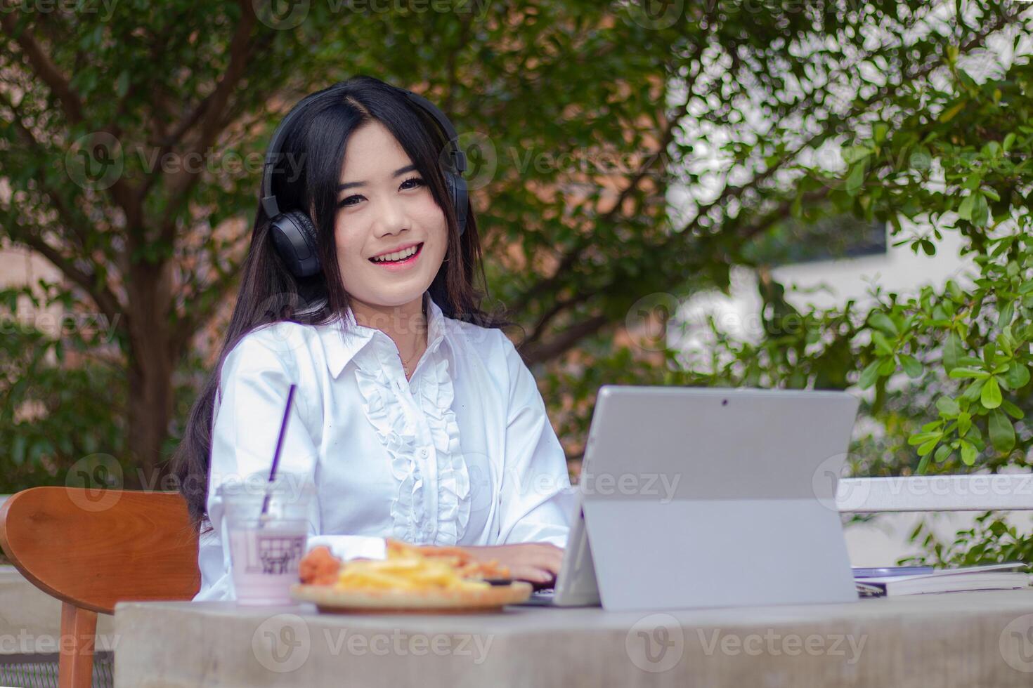 office beautiful asian woman in outdoor cafe. cheerful smiling girl listening to music while typing doing work in front of the laptop looking at the camera for content or promos photo