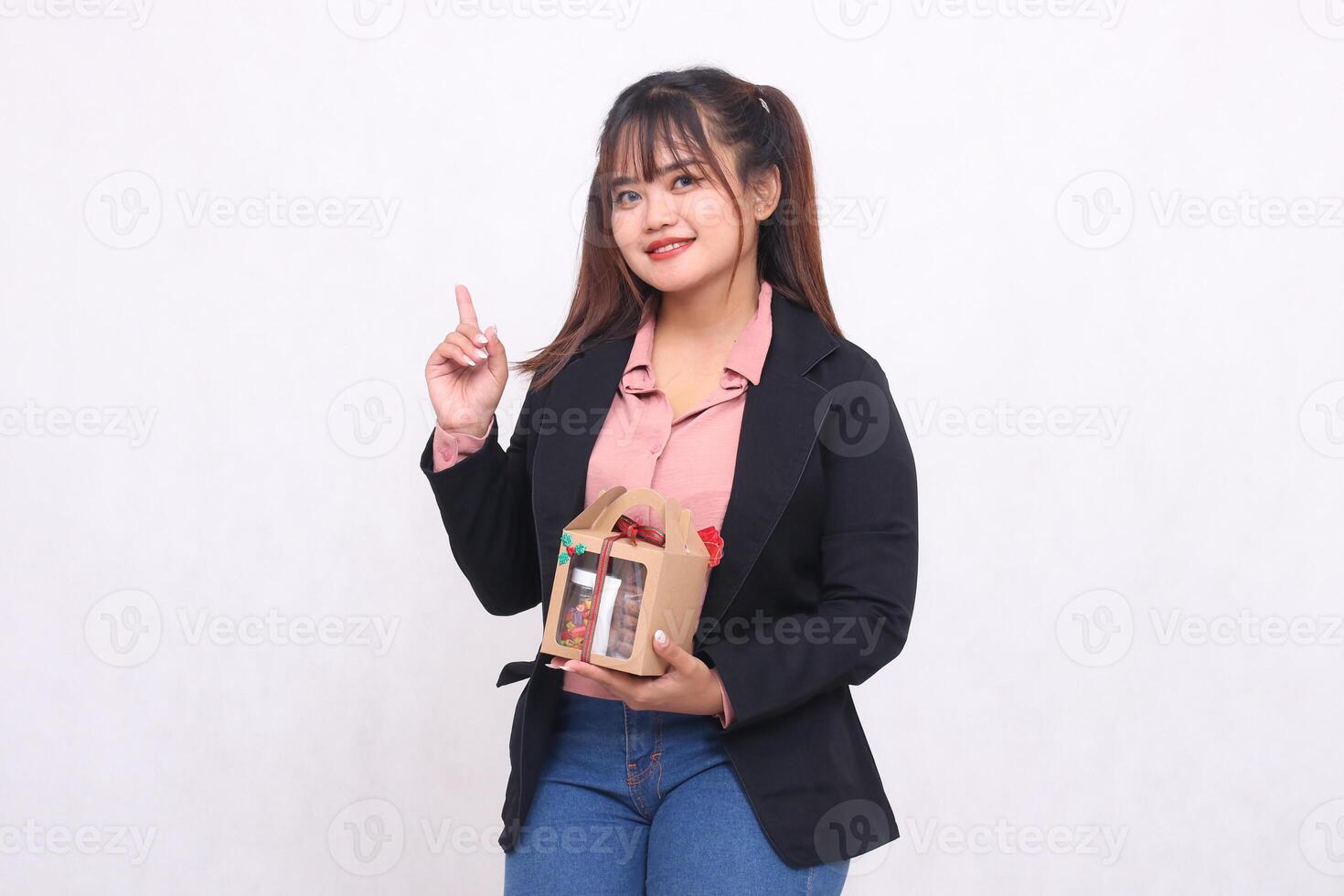 Beautiful Asian girl in her 20s smiles excitedly pointing up at Christmas gift in the form of hamper in hand on white background for advertising materials, banners, billboards photo