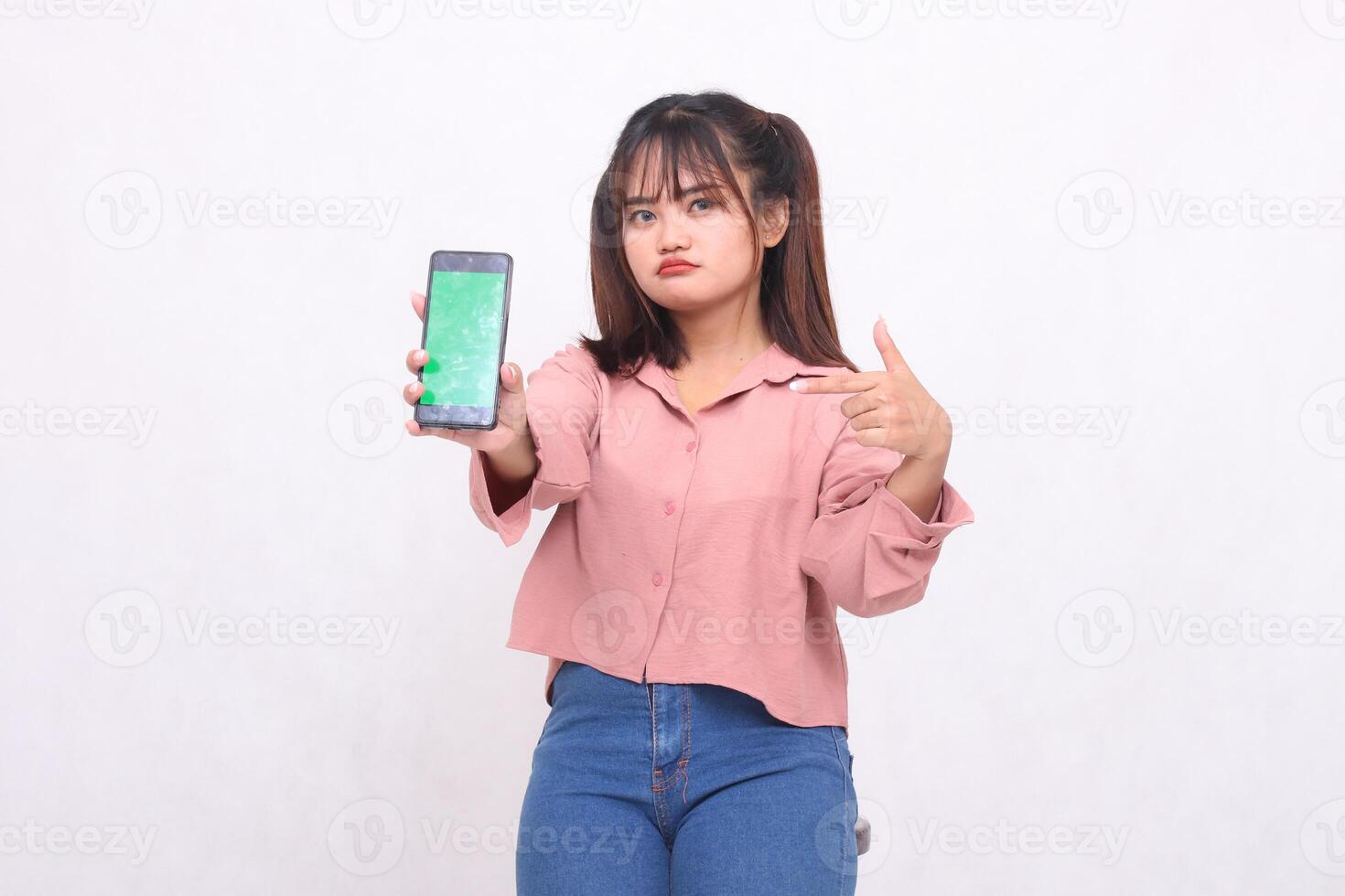 Beautiful happy Asian woman in her 20s wearing casual shirt holding white screen cellphone while frowning pointing at gadget on white background studio portrait for banner ad, banner, billboard photo