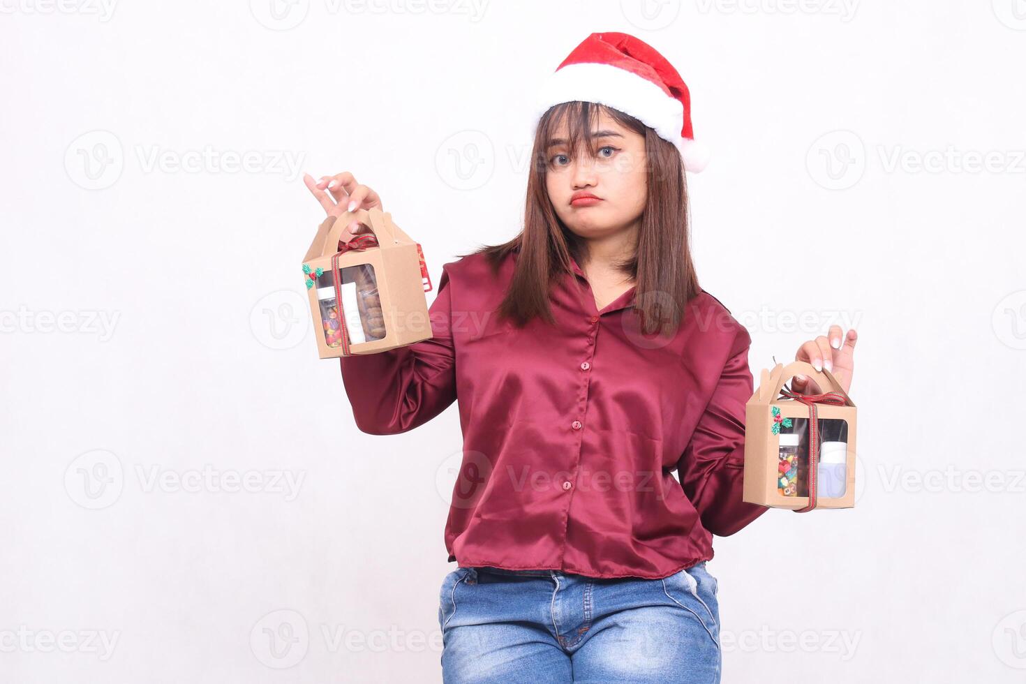 beautiful young southeast asian woman pouting at the camera carrying 2 boxes of hamper gifts at christmas wearing santa claus hat modern red shirt outfit white background for promotion and advertising photo