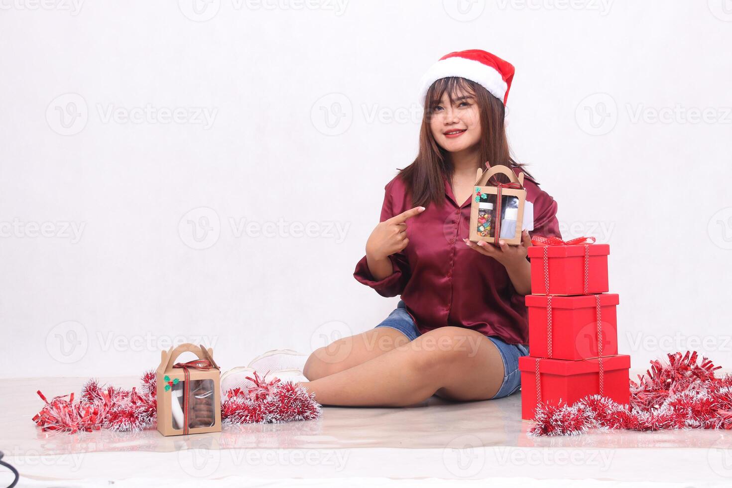 hermosa joven Sureste asiático niña sonriente señalando a cesto siguiente a 3 regalo cajas de cestas a Navidad vistiendo Papa Noel venda y rojo camisa en blanco antecedentes para promoción y publicidad foto