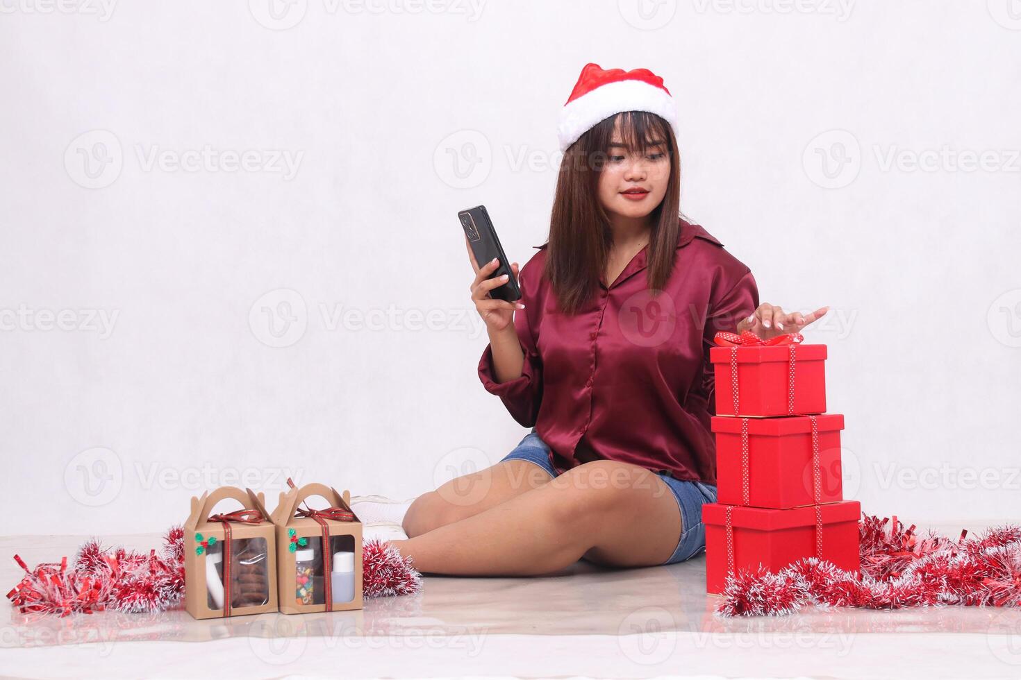 Beautiful young Southeast Asian girl checking orders next to 3 gift boxes of hampers at Christmas wearing Santa headband and red shirt on white background for promotion and advertising photo