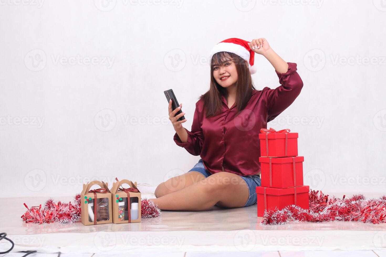 Beautiful young Southeast Asian girl successfully sent an order on a gadget next to 3 gift boxes of hampers at Christmas wearing a Santa headband and red shirt on a white background for promotion photo