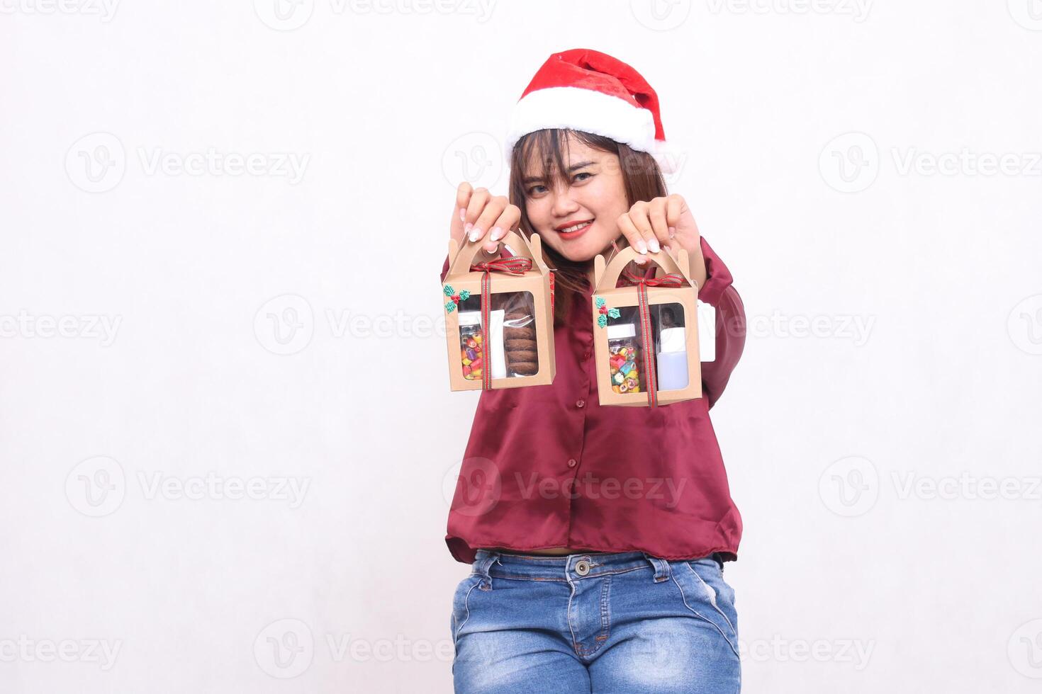 Beautiful young Southeast Asian woman smiling hands forward carrying 2 boxes of hamper gifts at Christmas wearing Santa Claus hat modern red shirt outfit white background for promotion and advertising photo