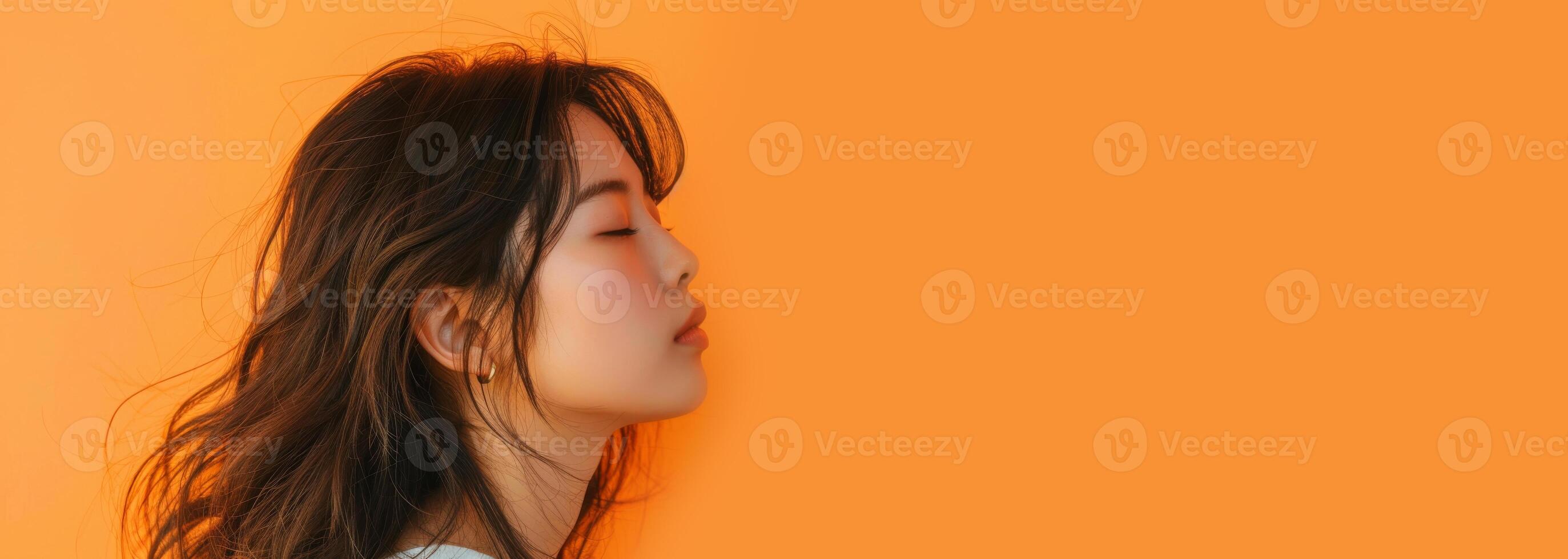 A woman with black hair stands in front of an orange wall, eyes closed, smiling photo