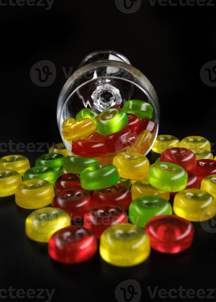 A lot of candy canes are scattered from an overturned glass goblet. Multi-colored candies in a glass on a black background. photo