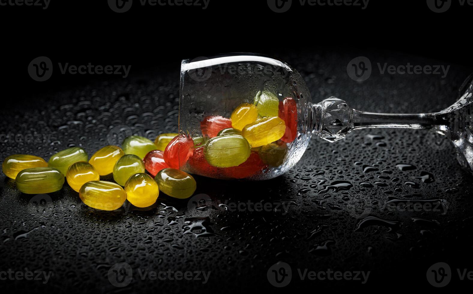 A lot of candy canes are scattered from an overturned glass goblet. Multi-colored candies in a glass on a black background. Marmalade candies. photo