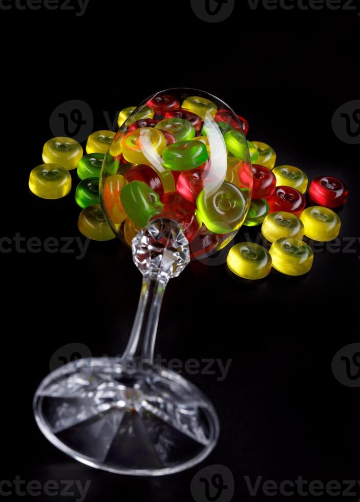 A lot of candy canes are scattered from an overturned glass goblet. Multi-colored candies in a glass on a black background. photo