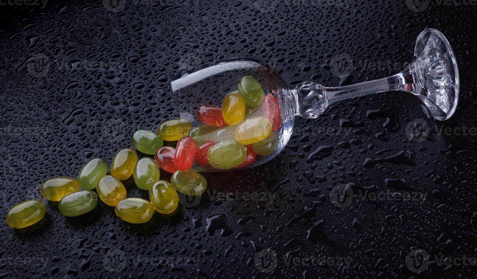 A lot of candy canes are scattered from an overturned glass goblet. Multi-colored candies in a glass on a black background. Marmalade candies. photo