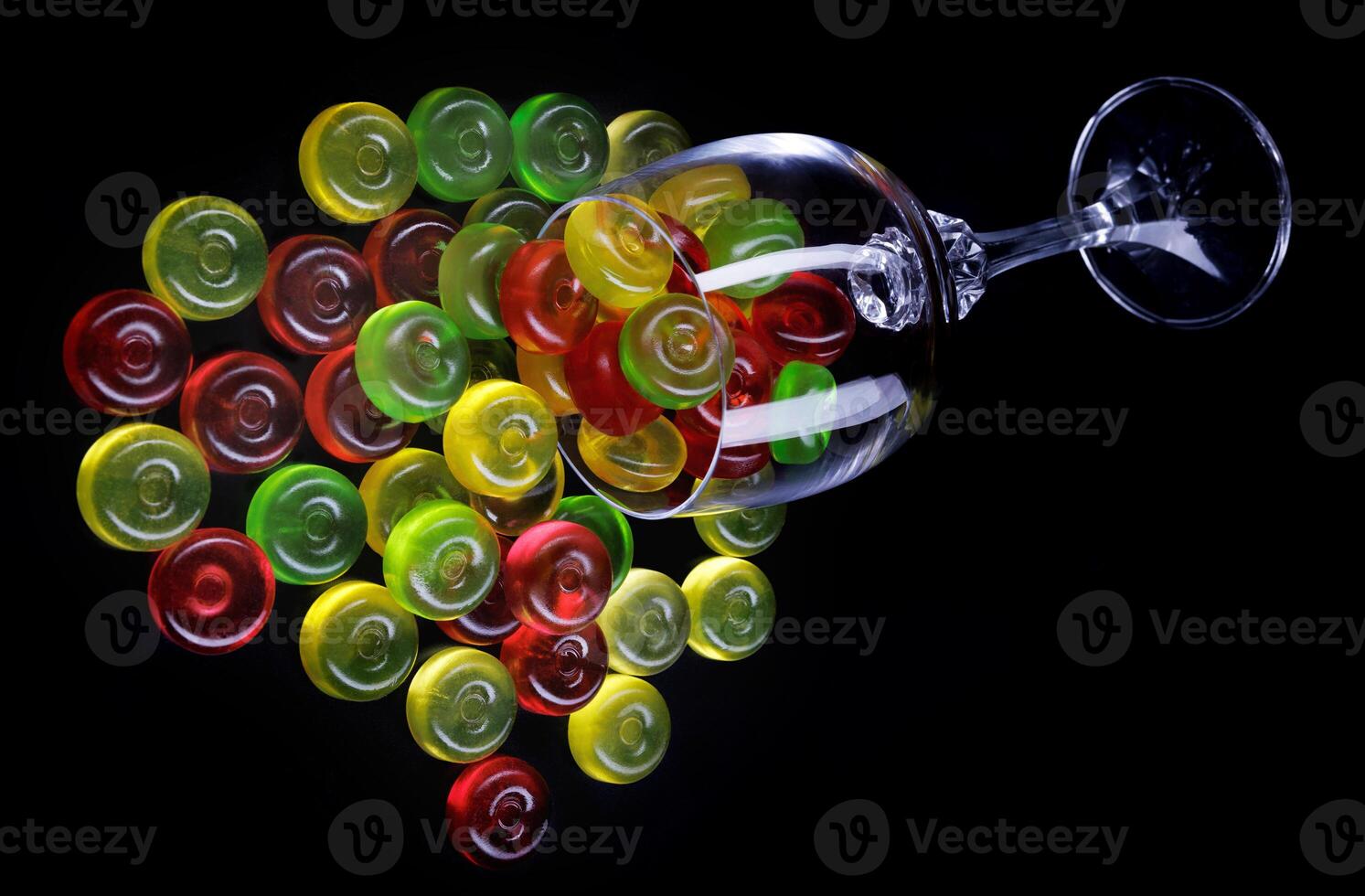 A lot of candy canes are scattered from an overturned glass goblet. Multi-colored candies in a glass on a black background. photo
