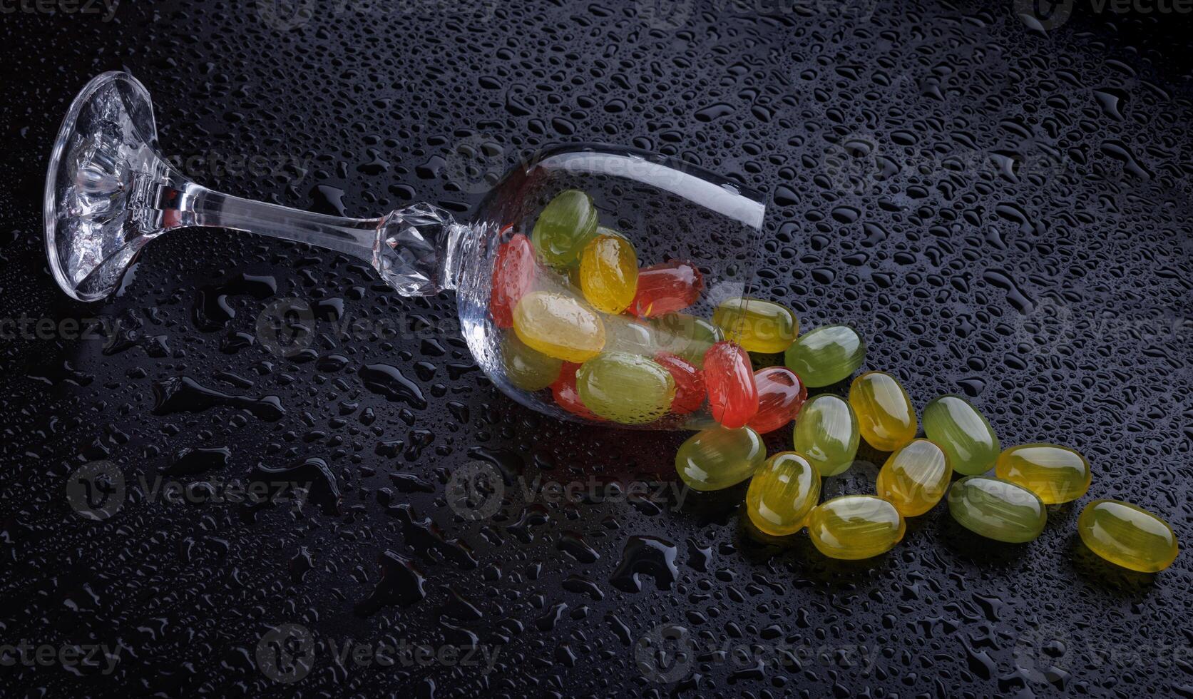 A lot of candy canes are scattered from an overturned glass goblet. Multi-colored candies in a glass on a black background. Marmalade candies. photo