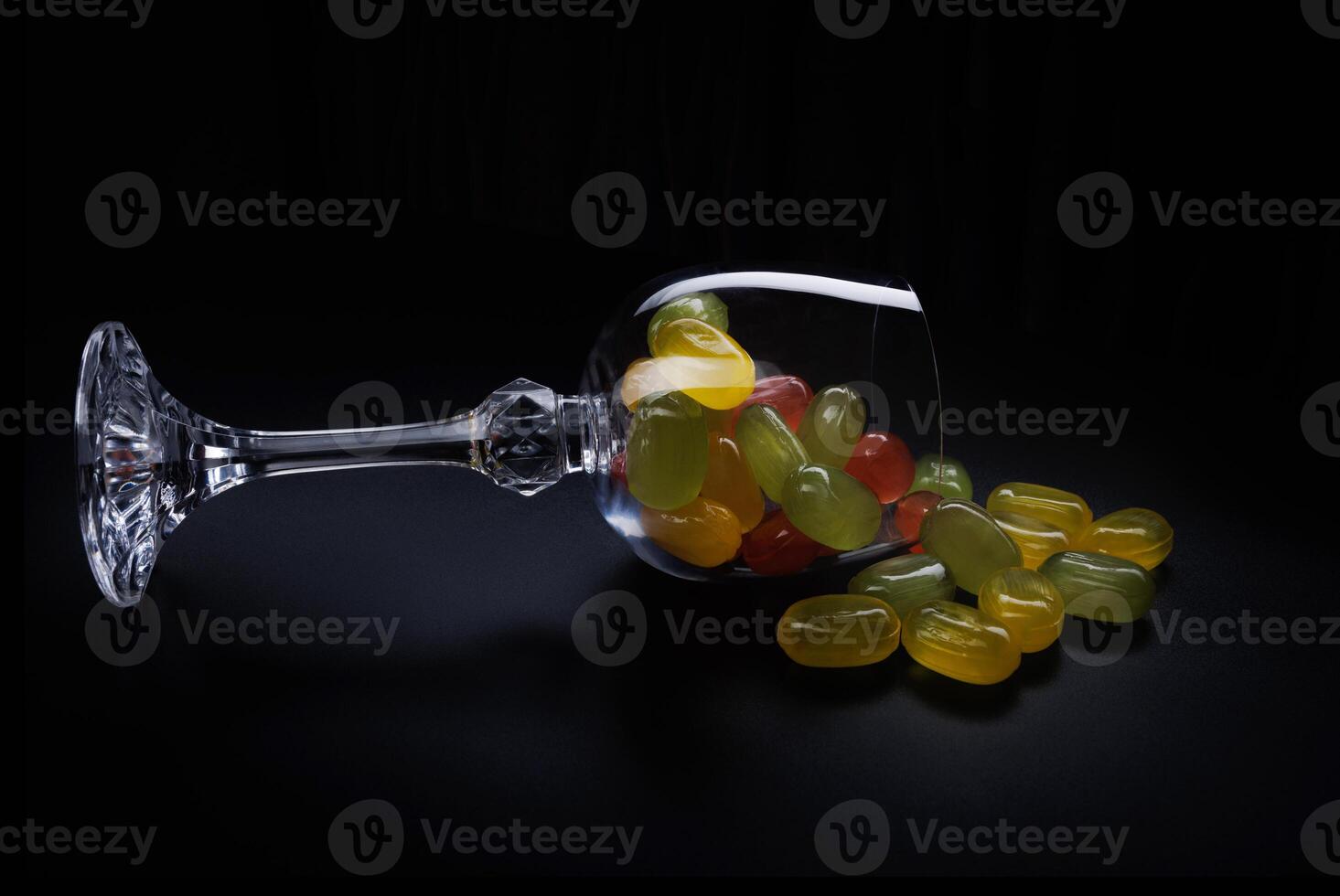 A lot of candy canes are scattered from an overturned glass goblet. Multi-colored candies in a glass on a black background. Marmalade candies. photo