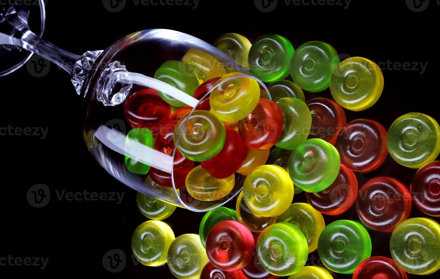 A lot of candy canes are scattered from an overturned glass goblet. Multi-colored candies in a glass on a black background. photo
