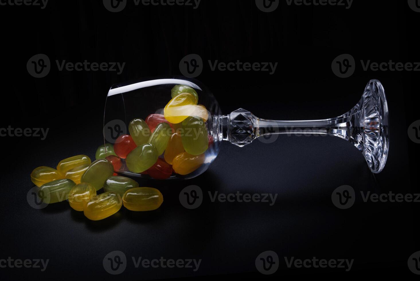 A lot of candy canes are scattered from an overturned glass goblet. Multi-colored candies in a glass on a black background. Marmalade candies. photo