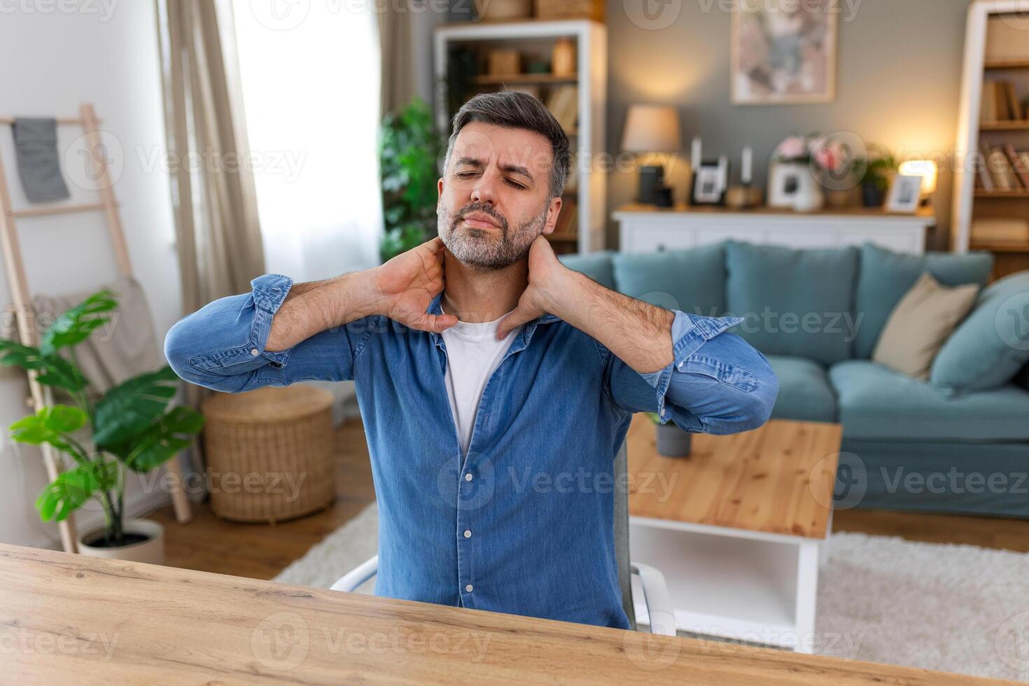 Businessman feeling pain in neck after sitting at the table with laptop. Tired man suffering of office syndrome because of long hours computer work. He is massaging his tense neck muscles photo