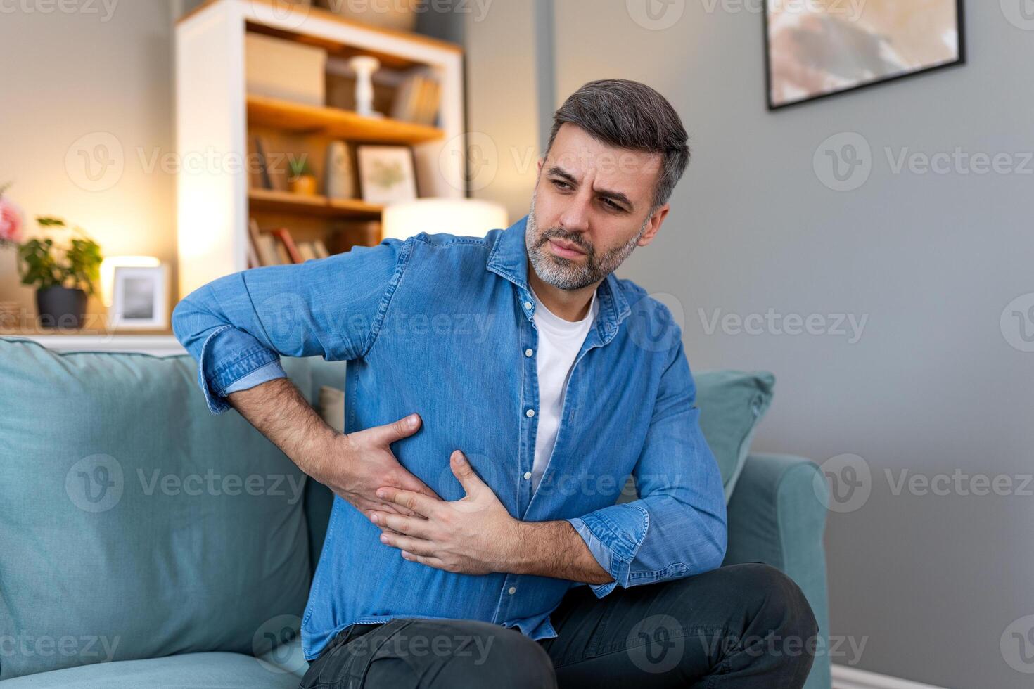 Terrible stomachache. Frustrated handsome young man hugging his belly and keeping eyes closed while lying on the couch at home photo
