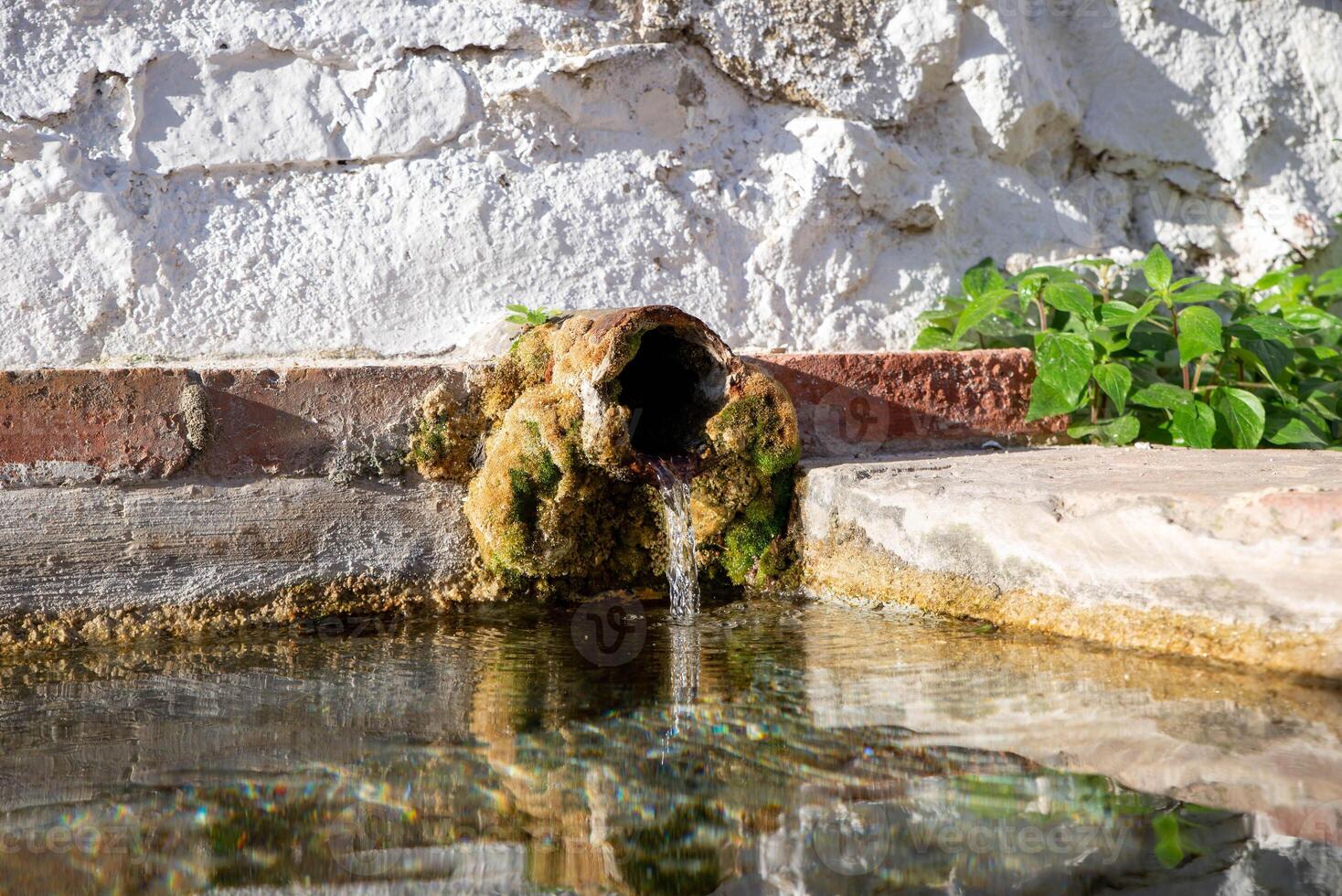 Clear water flows from a rusty, moss covered pipe protruding from a white plaster wall into a small pool. photo