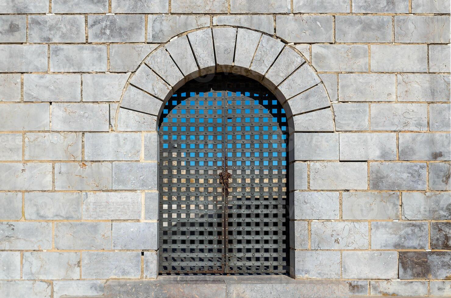 Rough gray old stone wall with grey iron gate closed with chain and padlock photo