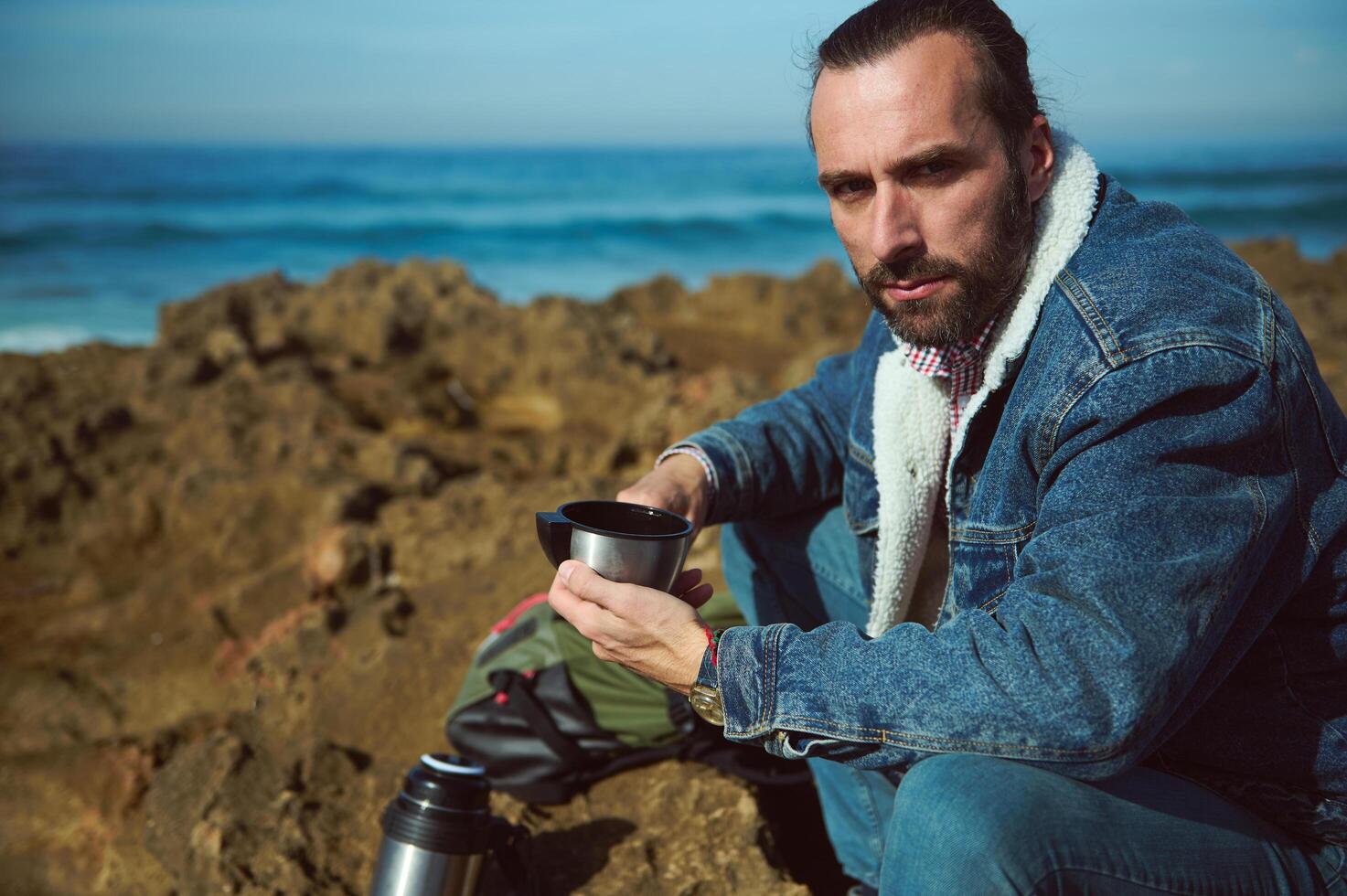 Lifestyle. Closeup view of a bearded man pouring hot water from a thermos, into a cup. The ocean in the background photo