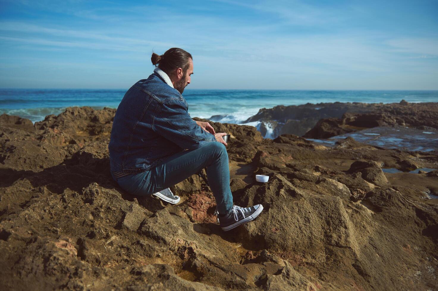 Active man resting on nature, sitting on a rocky beach and contemplating the ocean with breaking waves on the headland. photo