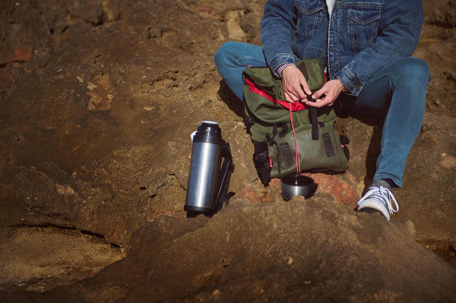 Close-up traveler with backpack and thermos flask, sitting on the cliff by sea, enjoying his coffee break on the nature photo