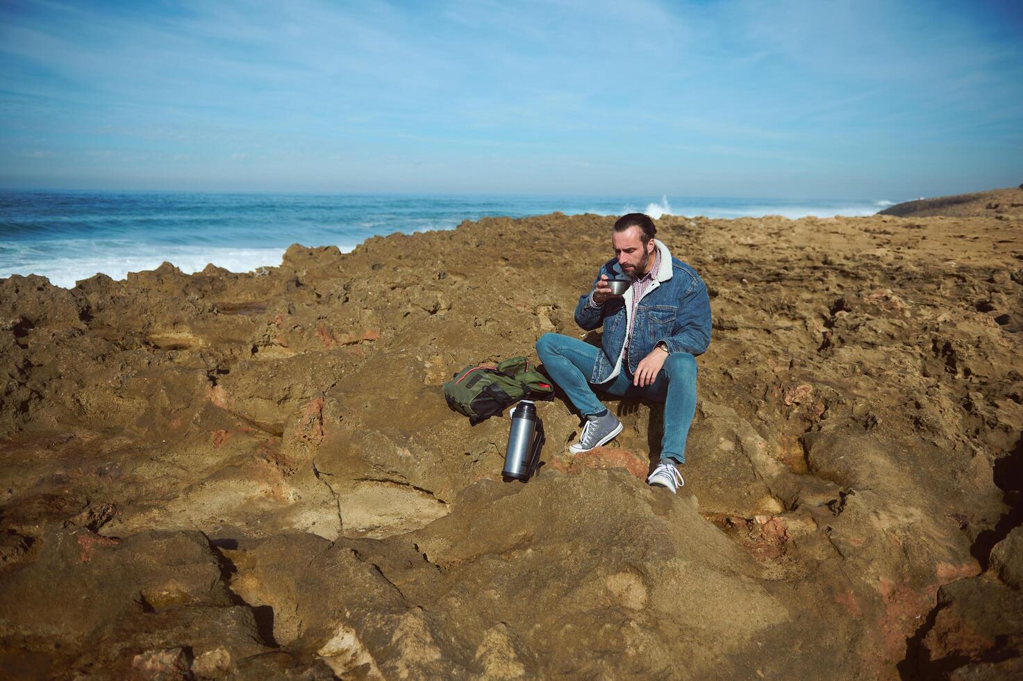 Full length portrait of a bearded man in blue denim, sitting on the rocky cliff during a coffee break. photo