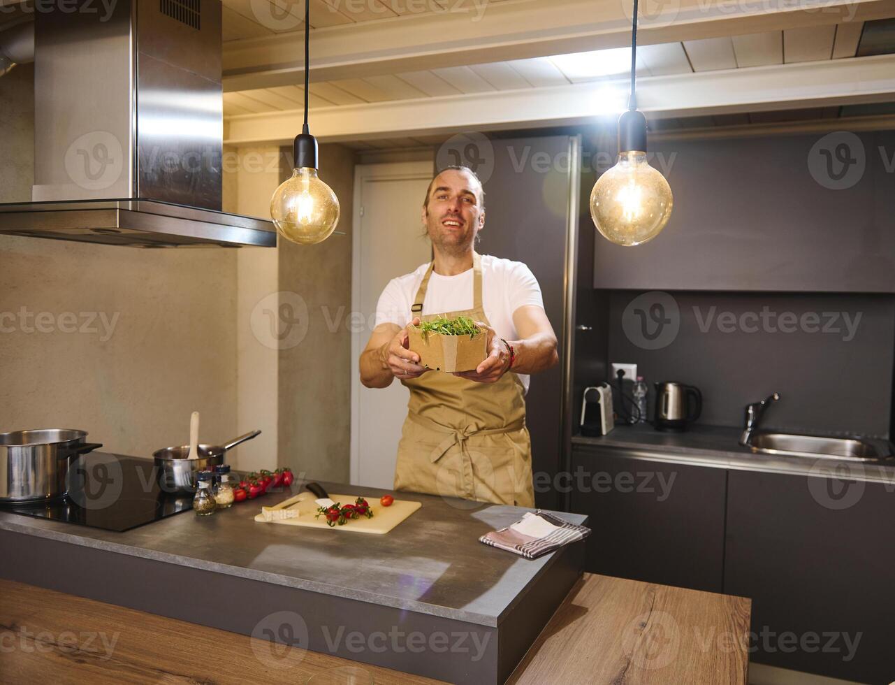 Handsome Caucasian young man chef smiles and holds out at camera a crate with arugula leaves, standing at kitchen countertop with fresh ingredients while cooking healthy dinner in the home kitchen. photo