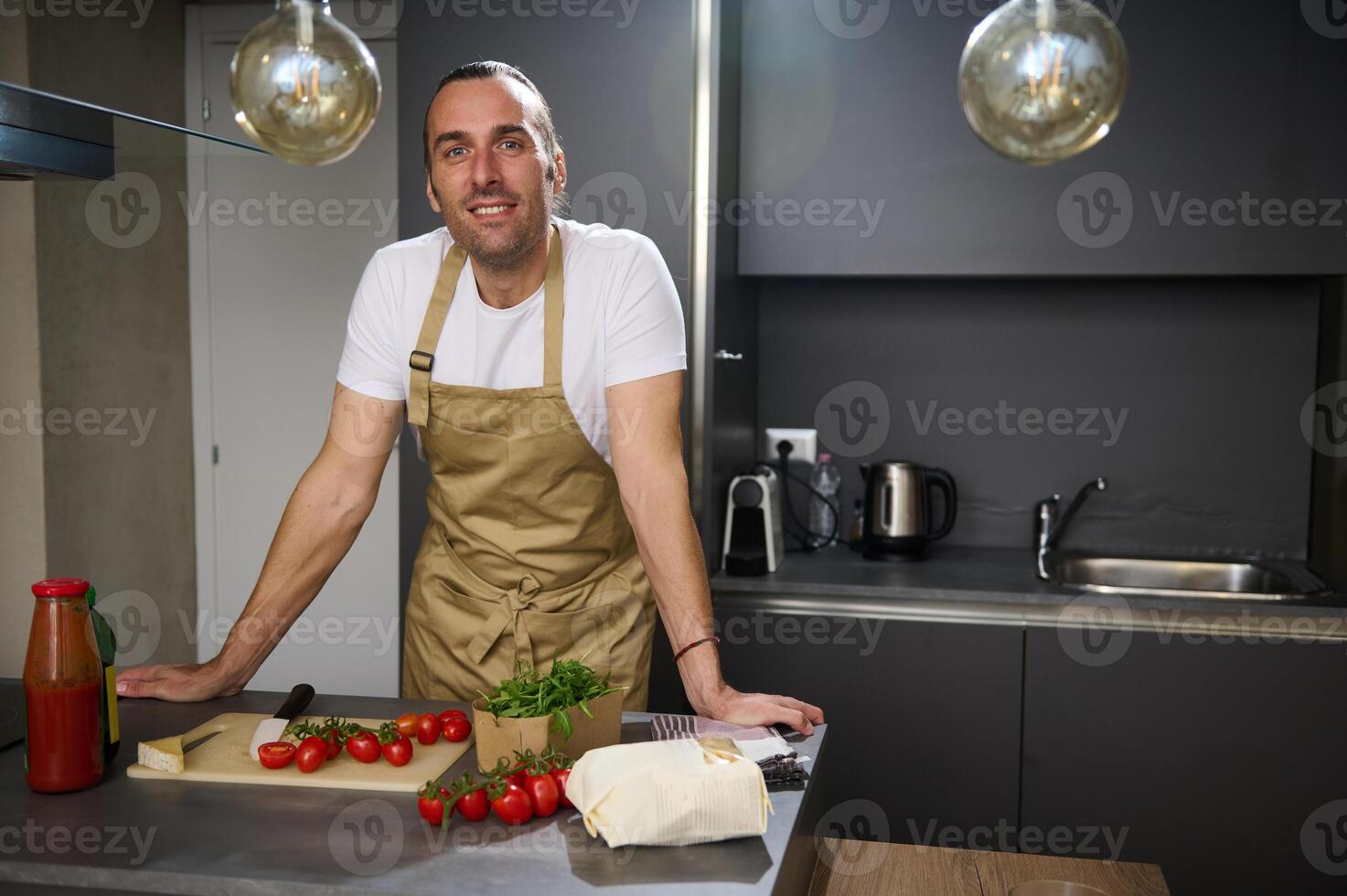 Authentic portrait of a handsome young adult man, a chef in beige apron, standing at kitchen counter with fresh ingredients, smiling looking confidently at camera, cooking healthy salad for dinner photo