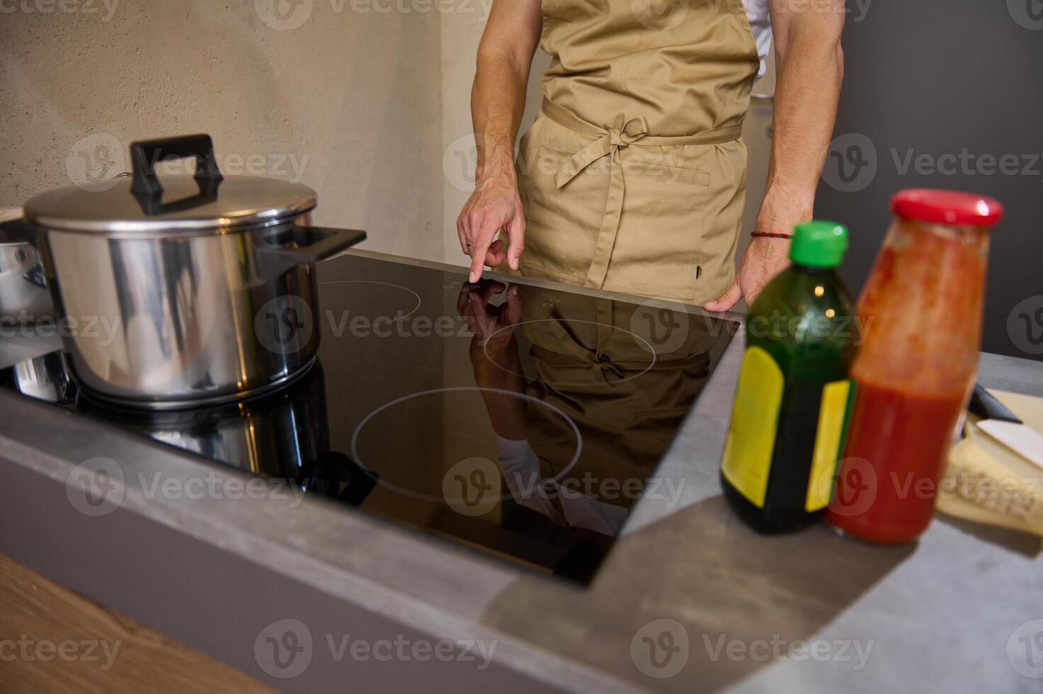 Close-up view of a man holding hand on the black glass surface of an electric or induction stove, switching it on, adjusting program while cooking dinner in a minimalist gray home kitchen interior photo