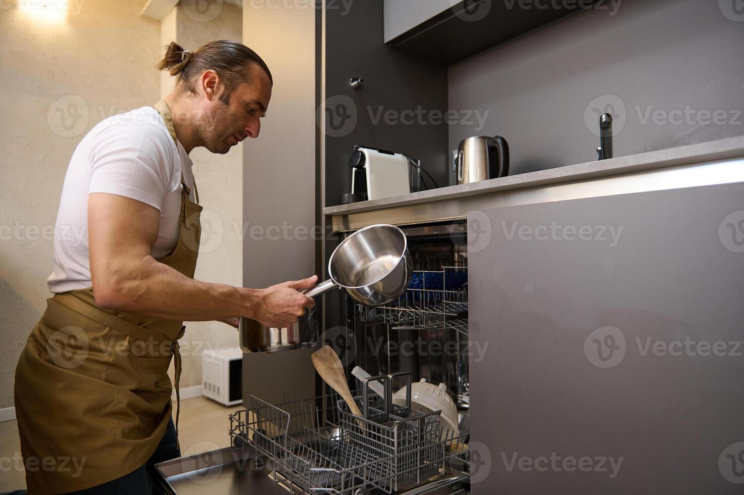 Handsome Caucasian young man in beige chef apron, takes out a clean saucepan from an overload automatic dishwasher with clean sterilize kitchen utensils. Modern kitchen appliances, lifestyle view photo
