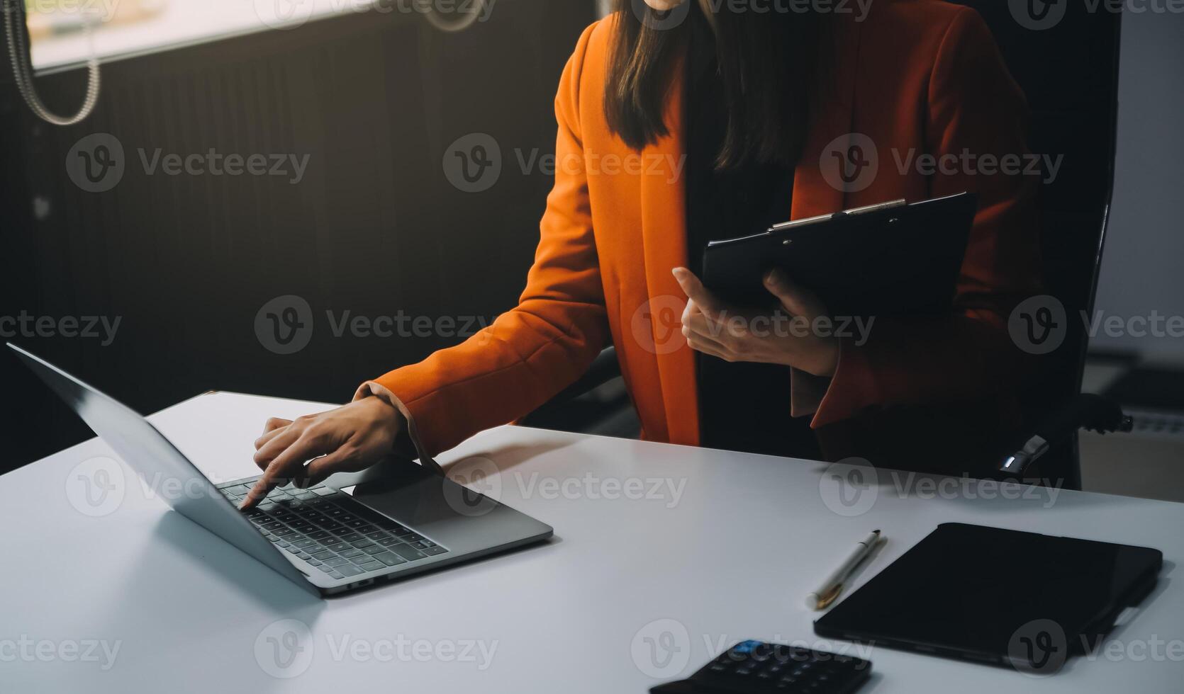 Smiling young Asian businesswoman working on a laptop computer at her desk in a bright modern home office, doing calculating expense financial report finance making notes on paper graph data document. photo