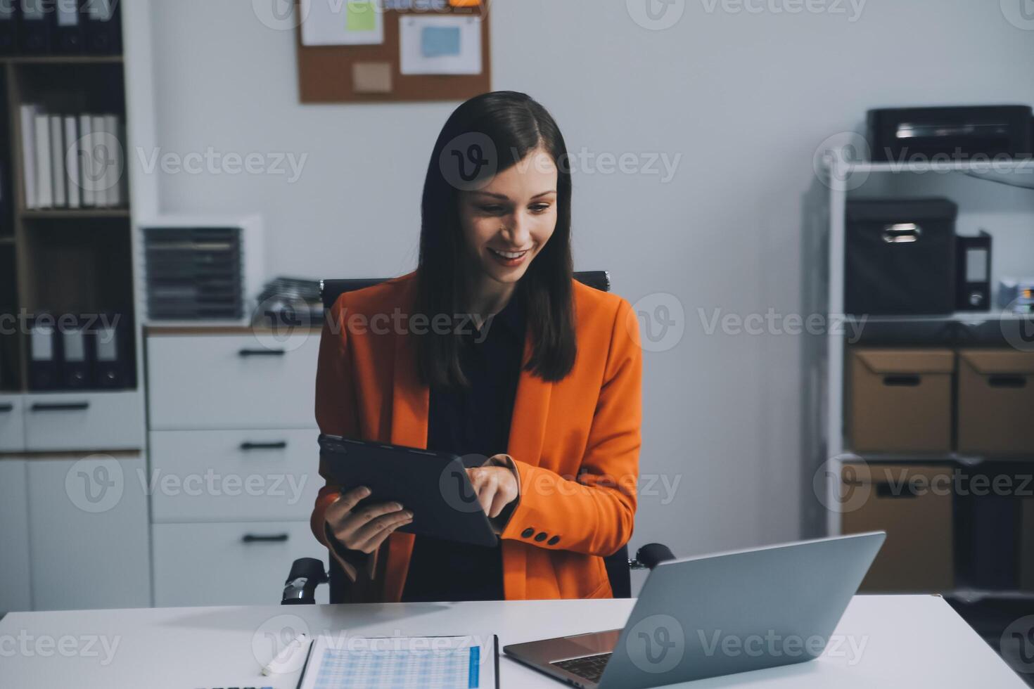 Smiling young Asian businesswoman working on a laptop computer at her desk in a bright modern home office, doing calculating expense financial report finance making notes on paper graph data document. photo