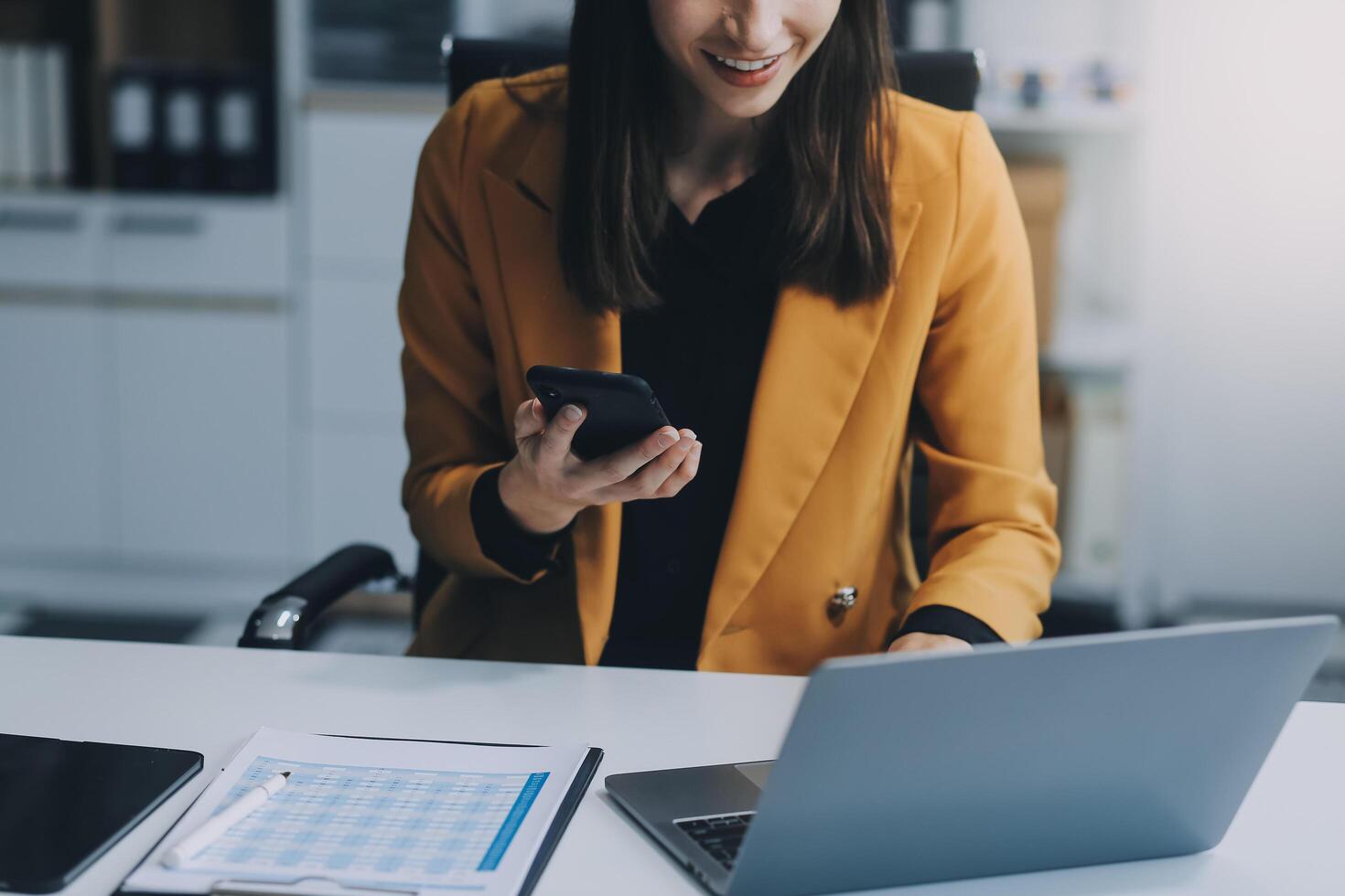 Beautiful Asian woman using a smartphone and working with a laptop while sitting at an office desk, working from home concept. photo
