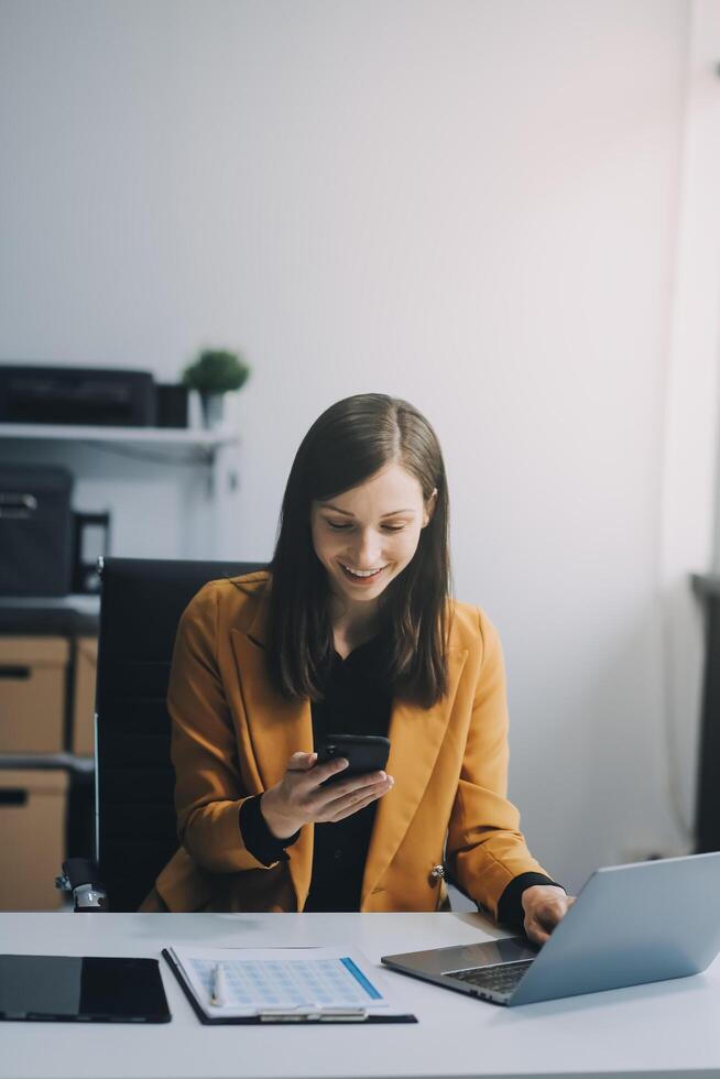 Beautiful Asian woman using a smartphone and working with a laptop while sitting at an office desk, working from home concept. photo