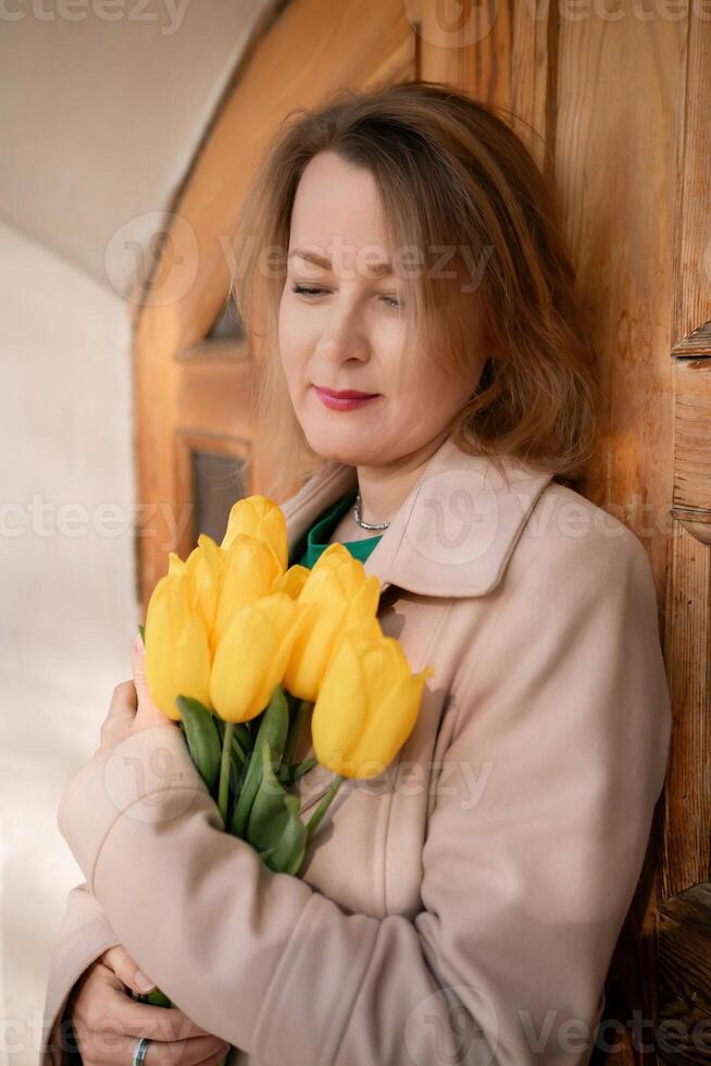 A woman is holding a bouquet of yellow tulips. She is wearing a tan coat and a green shirt. photo