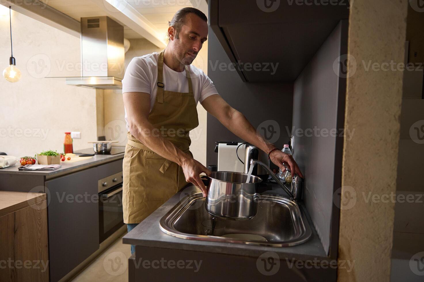 Attractive handsome Caucasian young man in beige chef's apron, filling a stainless steel saucepan with running water until it boils and cooks something in a minimalist home kitchen interior photo