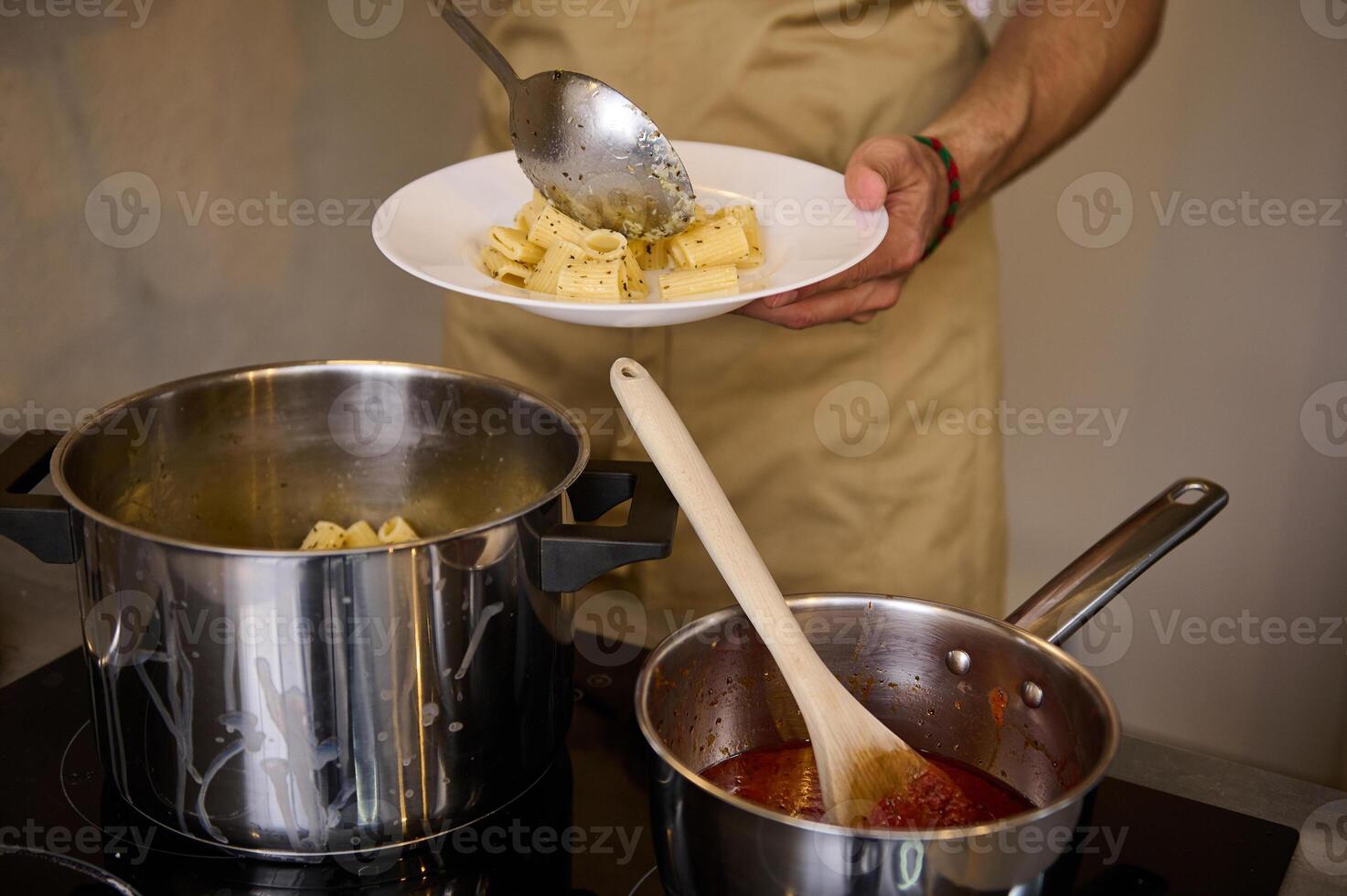 Close-up male chef plating up freshly cooked pasta, putting boiled penne on a white plate, standing at induction electric stove with saucepan full of tomato pasta. Italian culinary. Food background photo