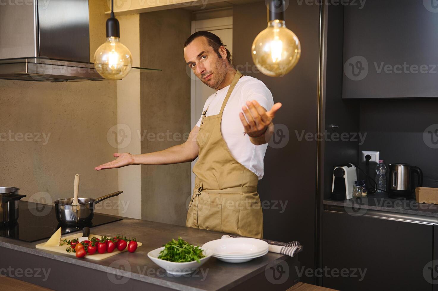 Handsome Caucasian man in chef's apron cooking family dinner, smiling and showing the kitchen counter with fresh ingredients looking at camera photo