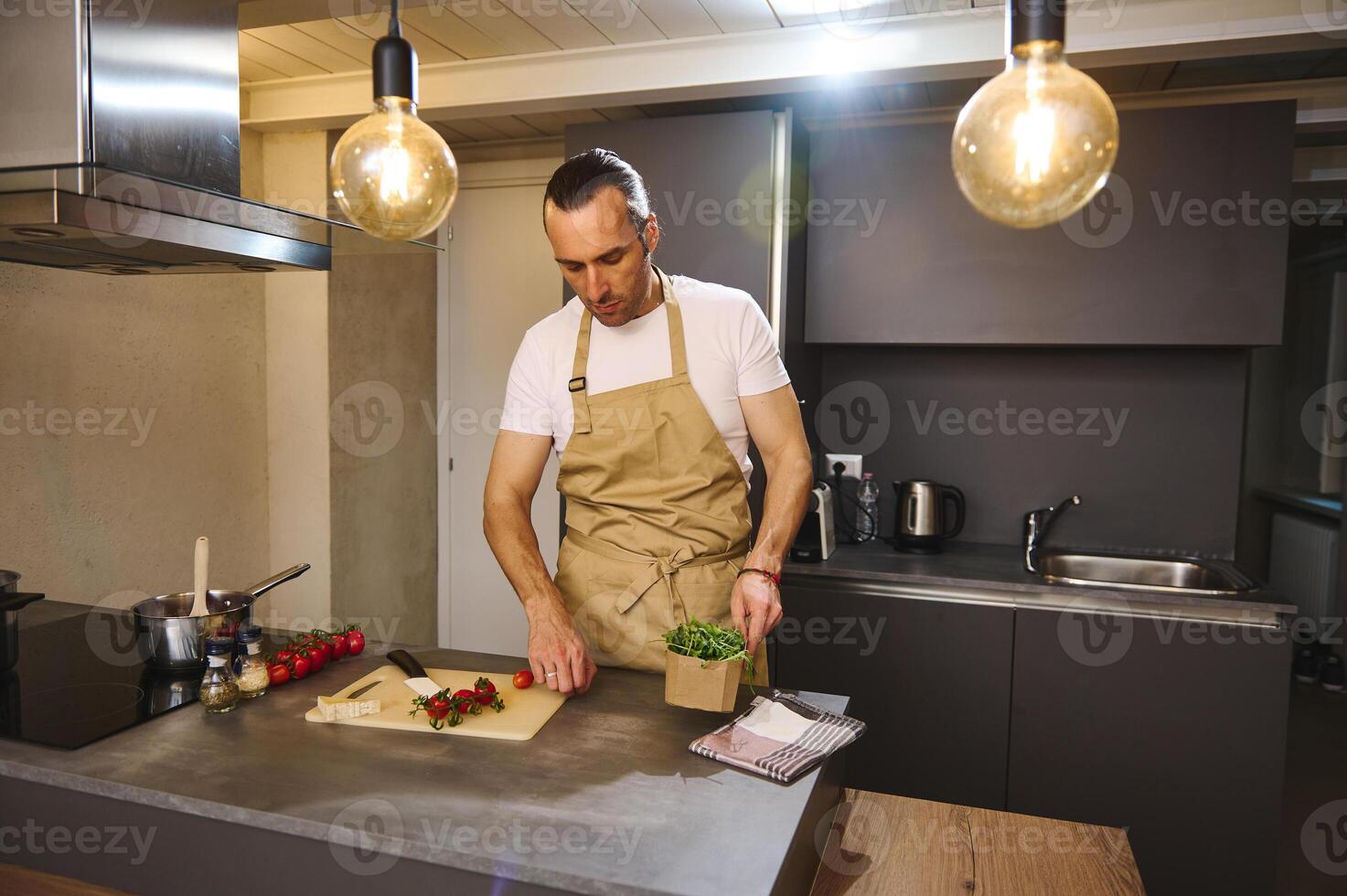 Handsome young adult man 40s, dressed in beige chef apron holding a cardboard crate with fresh green leaves of arugula, cooking healthy dinner in the home kitchen. photo