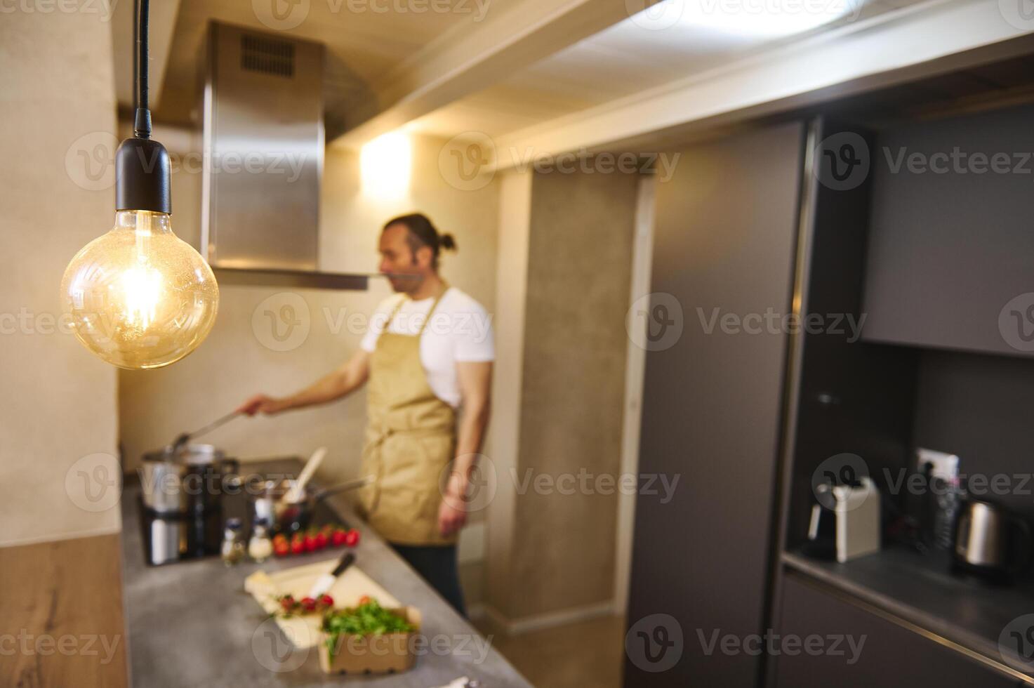 Details on a stylish vintage lighted lamp in modern kitchen interior, against blurred male chef standing at electric stove and stirring ingredients, preparing Italian traditional tomato sauce photo