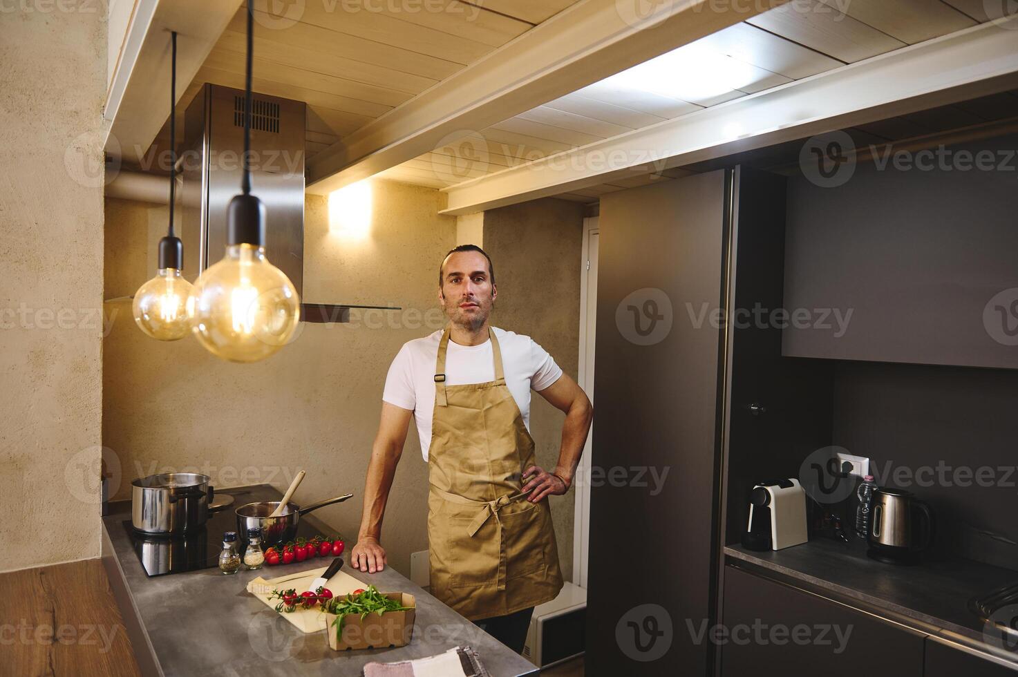 Authentic portrait of a handsome young adult man in beige chef's apron, standing at kitchen counter and looking at camera while cooking dinner, in the modern interior of minimalist home kitchen photo