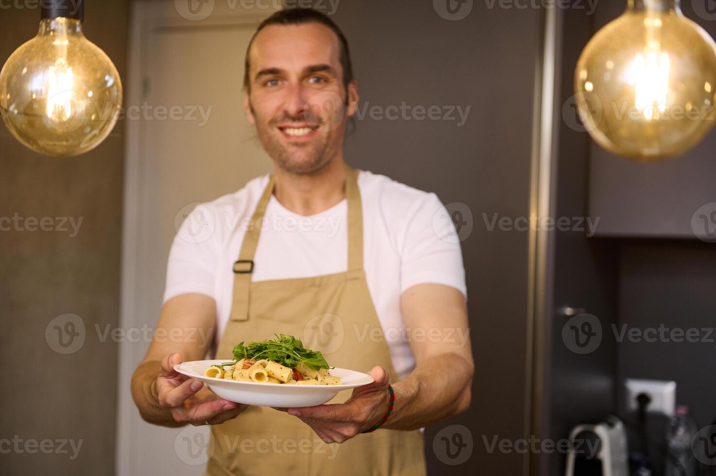 Authentic portrait of a smiling Caucasian man in beige chef's apron, holding a plate with Italian spaghetti pasta and showing it at camera. standing modern home kitchen interior. People and culinary photo