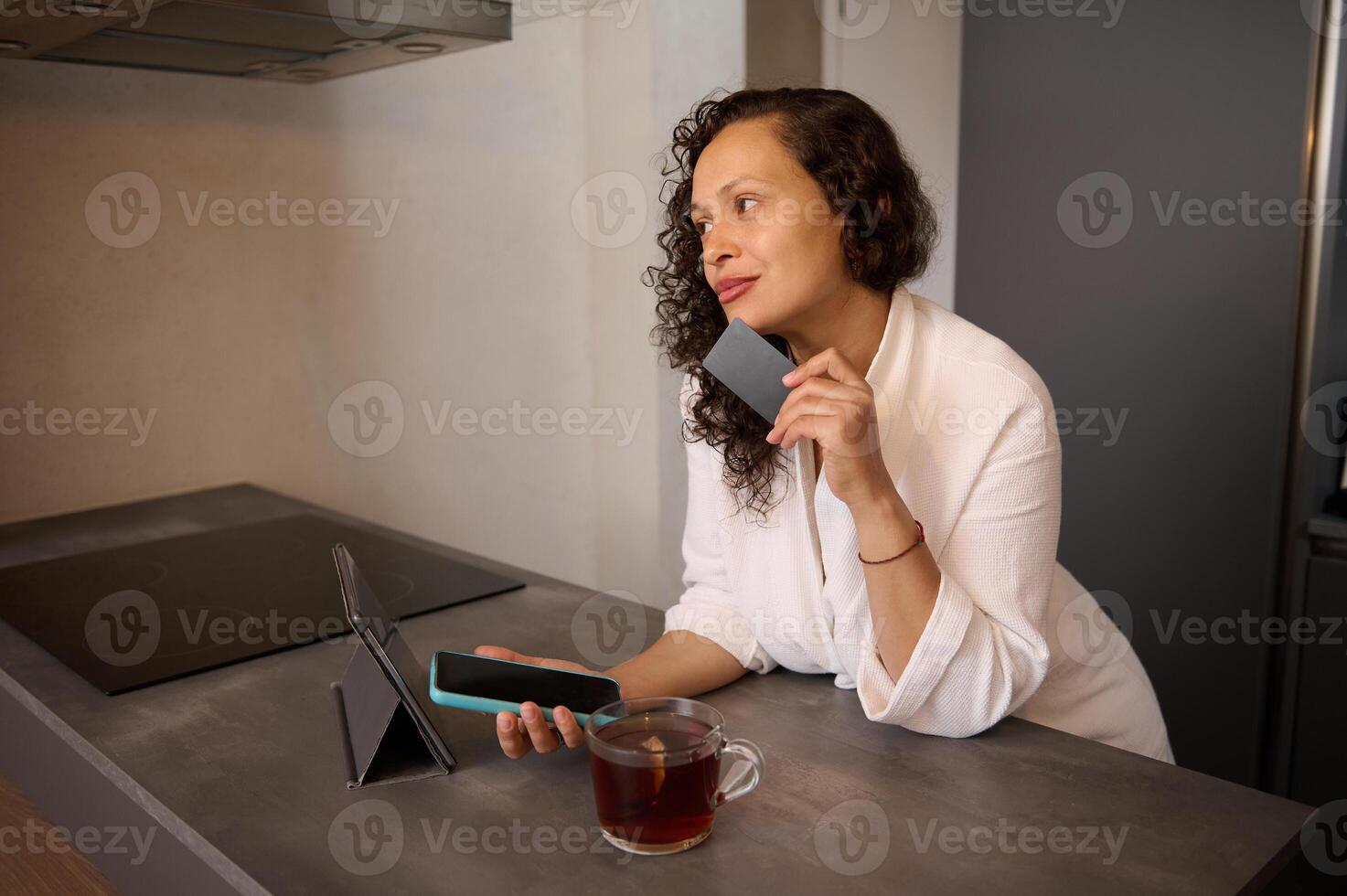 Young adult pretty woman in white bathrobe at home, making online payments via internet mobile banking, sitting at table with smart phone and credit card in her hands photo