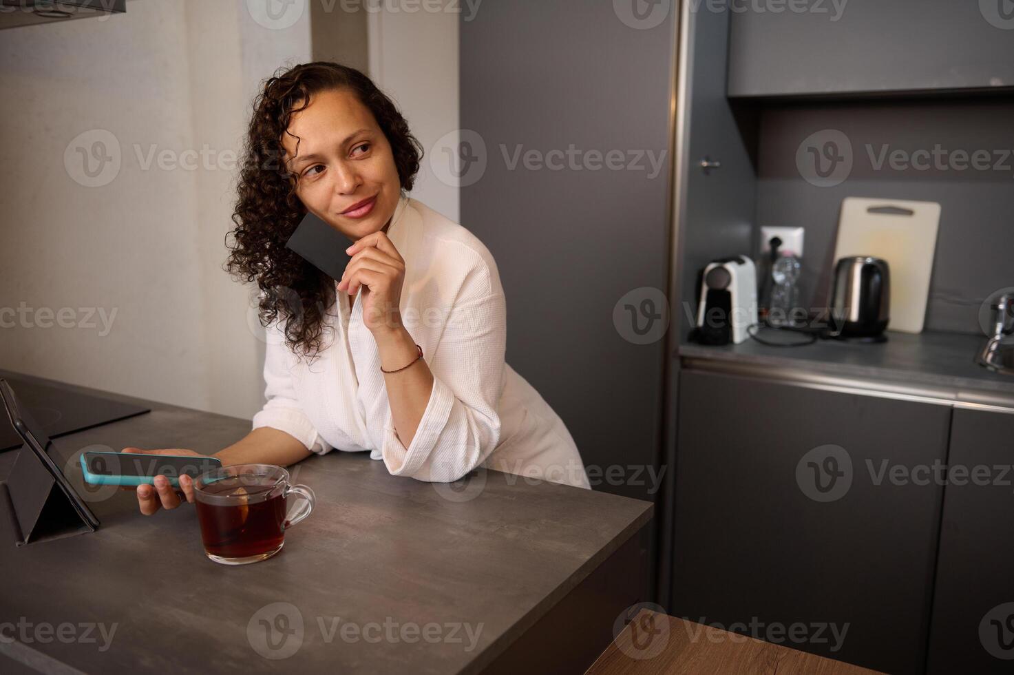Middle aged confident beautiful woman holding bank credit card, using smartphone, online shopping, paying bills, receiving or making payment via mobile internet banking, standing at table at home photo