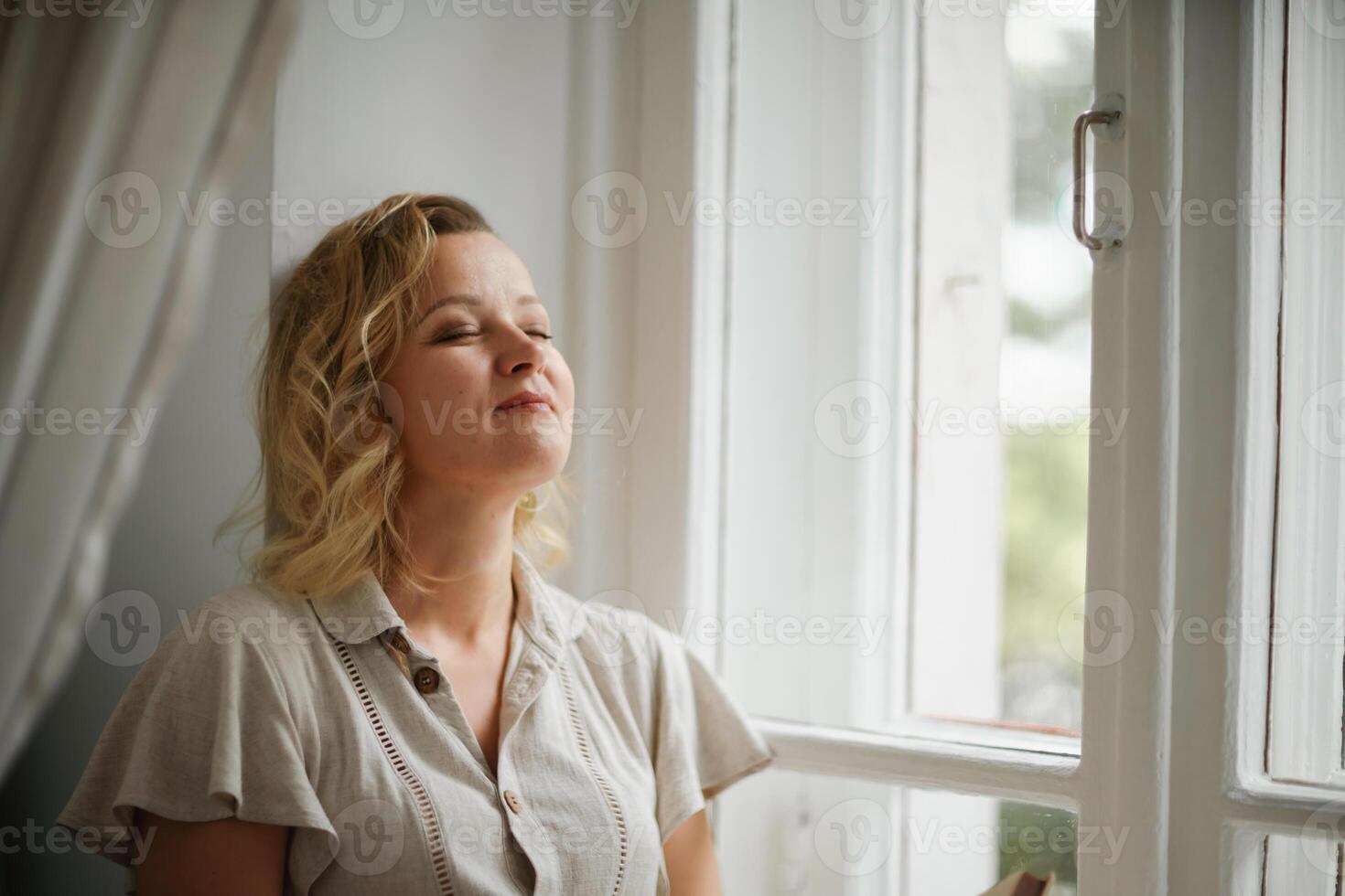 A middle-aged woman in a cream dress sits mysteriously and looks out the window on the windowsill. Green trees outside. photo